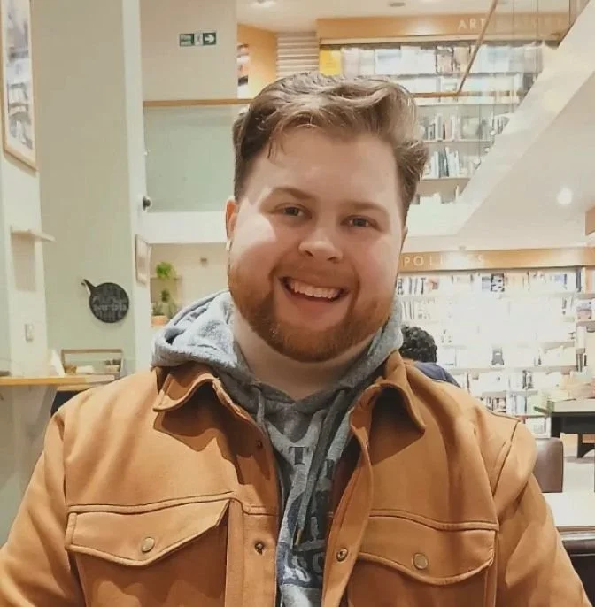 A smiling man with a beard and short hair, wearing a tan jacket over a gray hoodie, sitting in a bookstore or library.