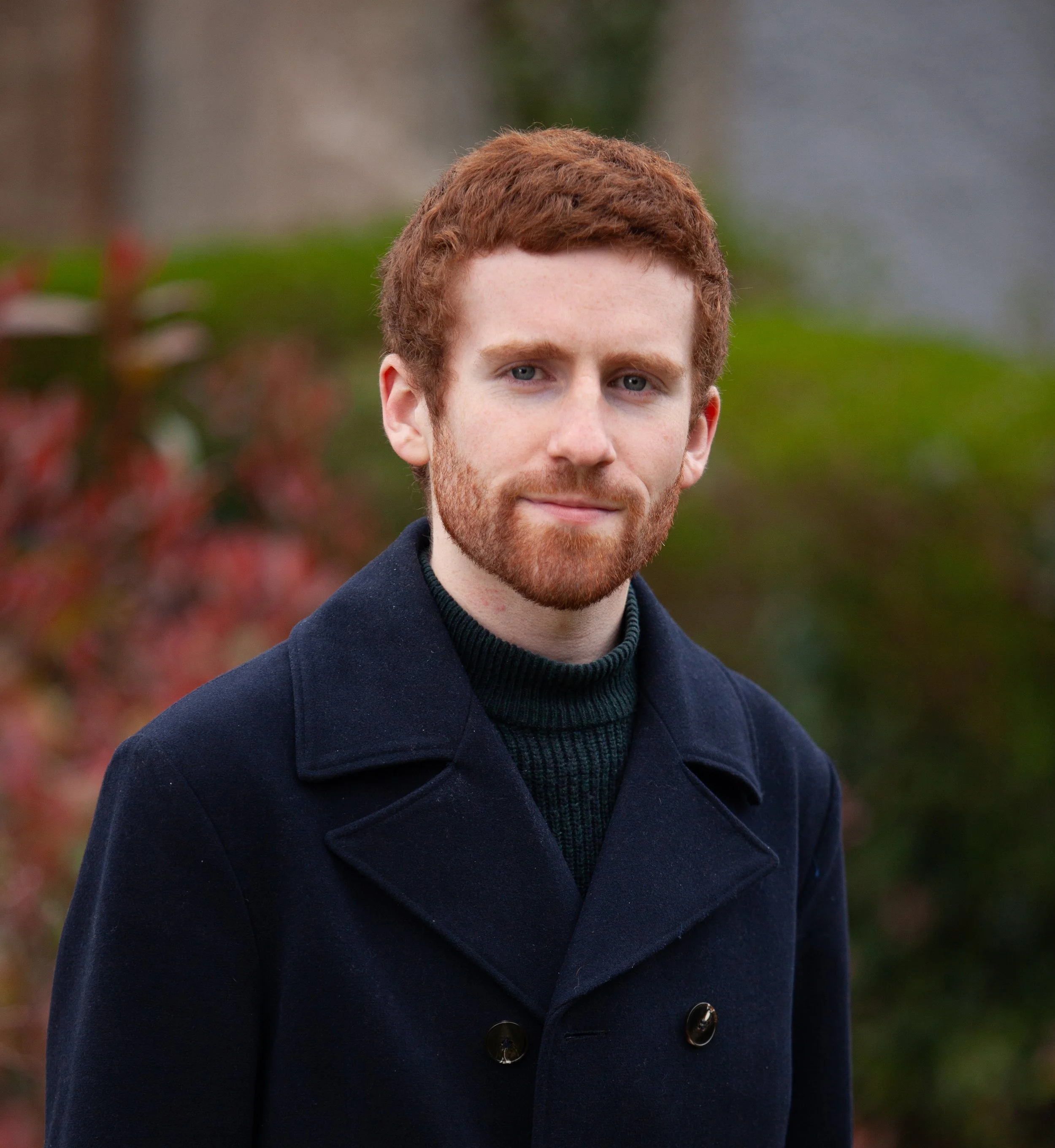 A young man with red hair and beard wearing a navy peacoat stands outdoors with blurred autumnal background.