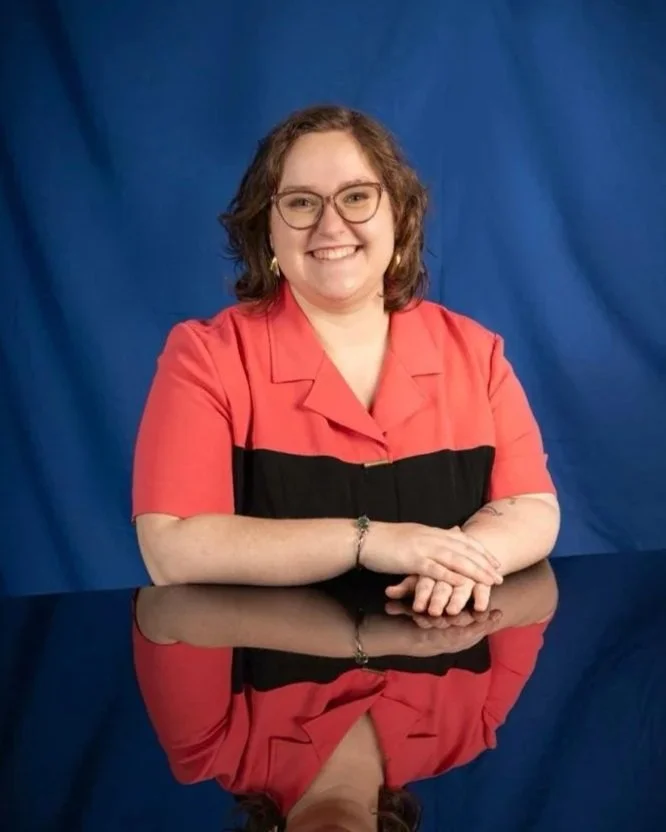 A woman with curly brown hair, glasses, and a red and black shirt, sitting at a reflective black table with a blue background.