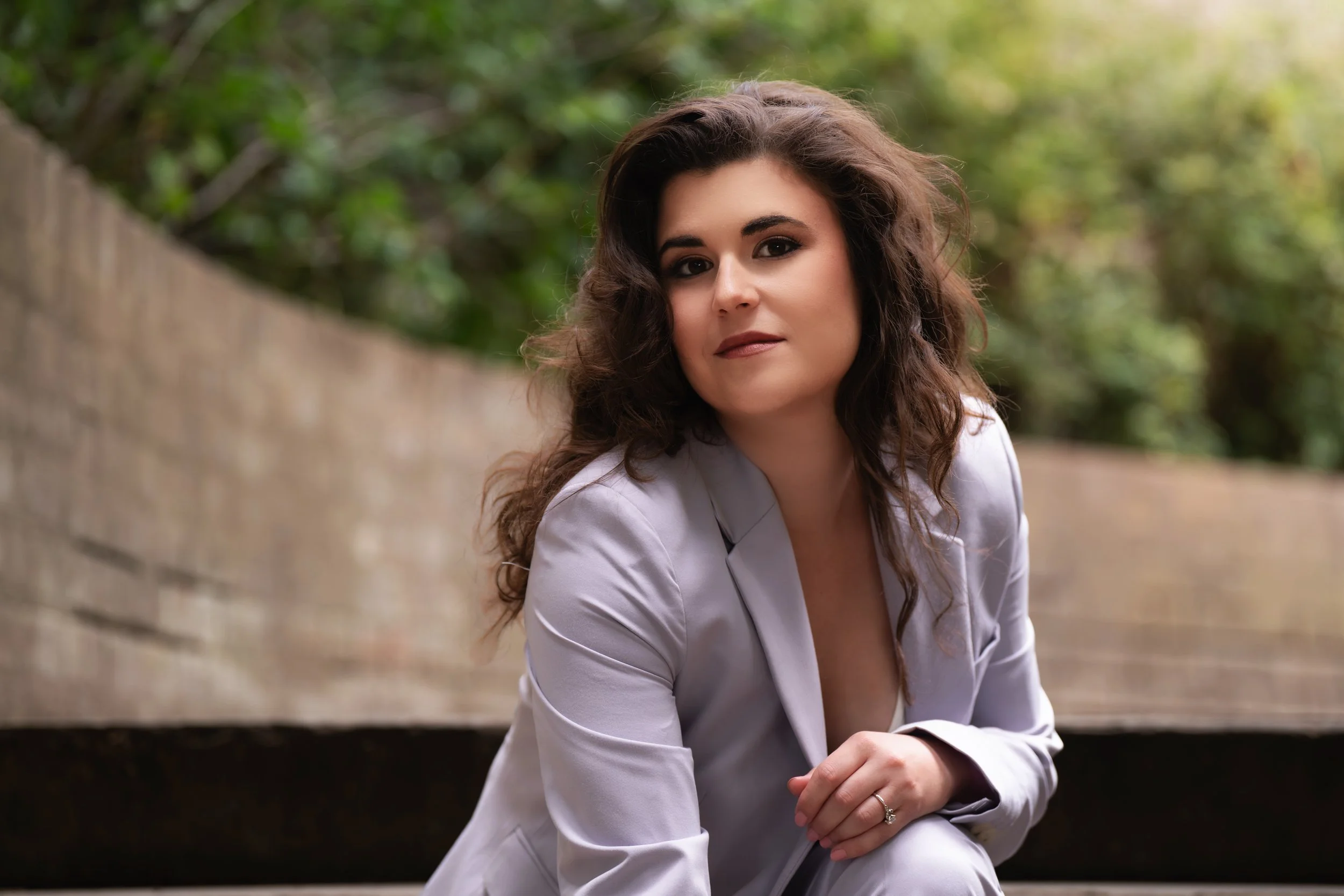 A woman with dark, wavy hair in a light gray blazer, sitting outdoors on steps with a brick background and green foliage, looking at the camera.