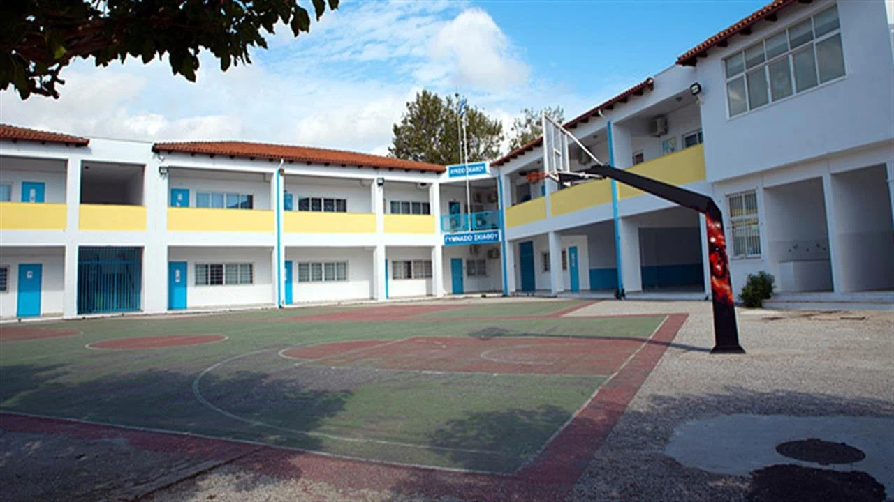 An outdoor basketball court surrounded by a white two-story school building with blue and yellow accents, under a partly cloudy sky.