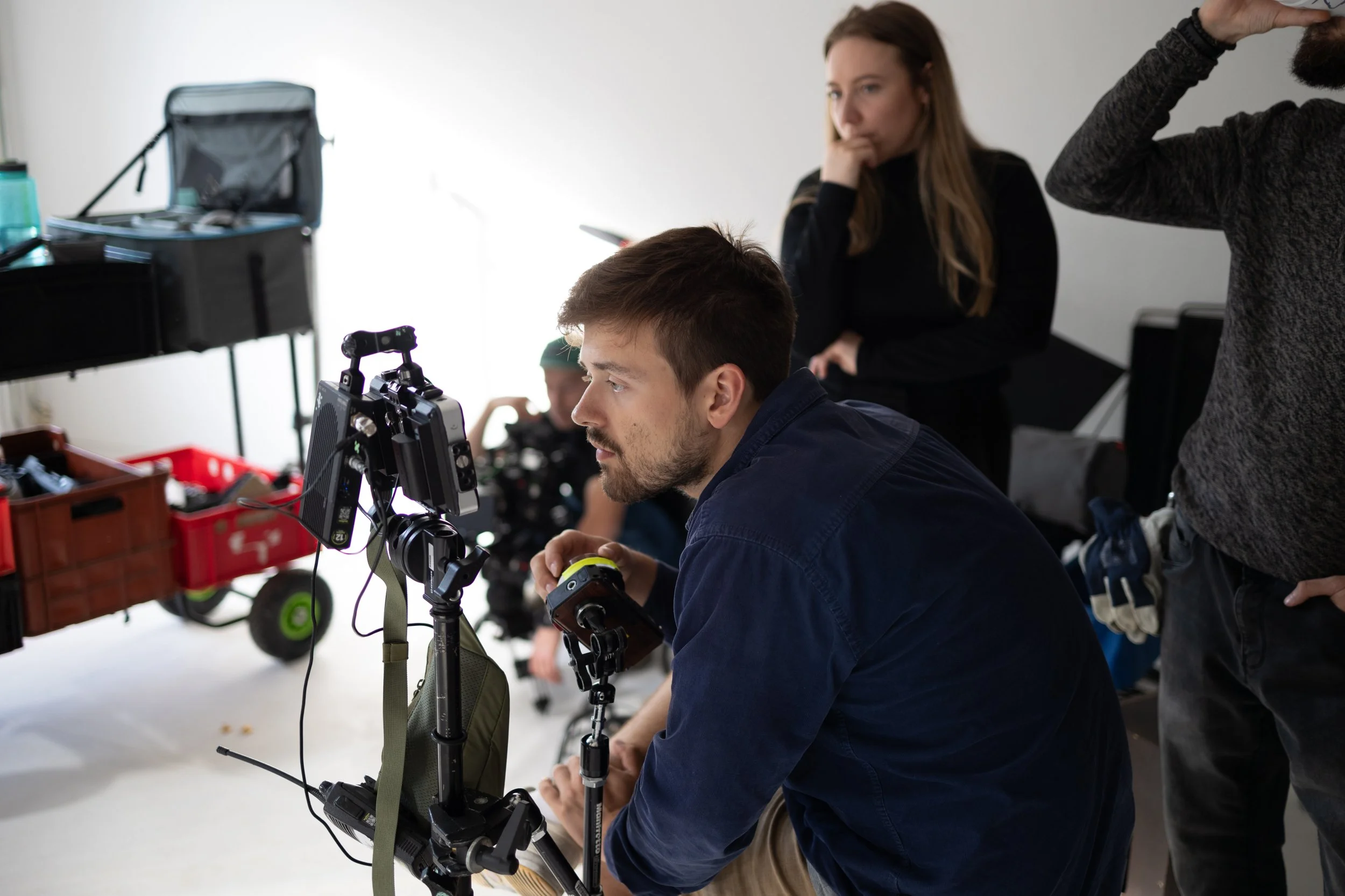 A group of people on a film set, with a man operating a camera in the foreground, a woman watching attentively, and others working in the background.