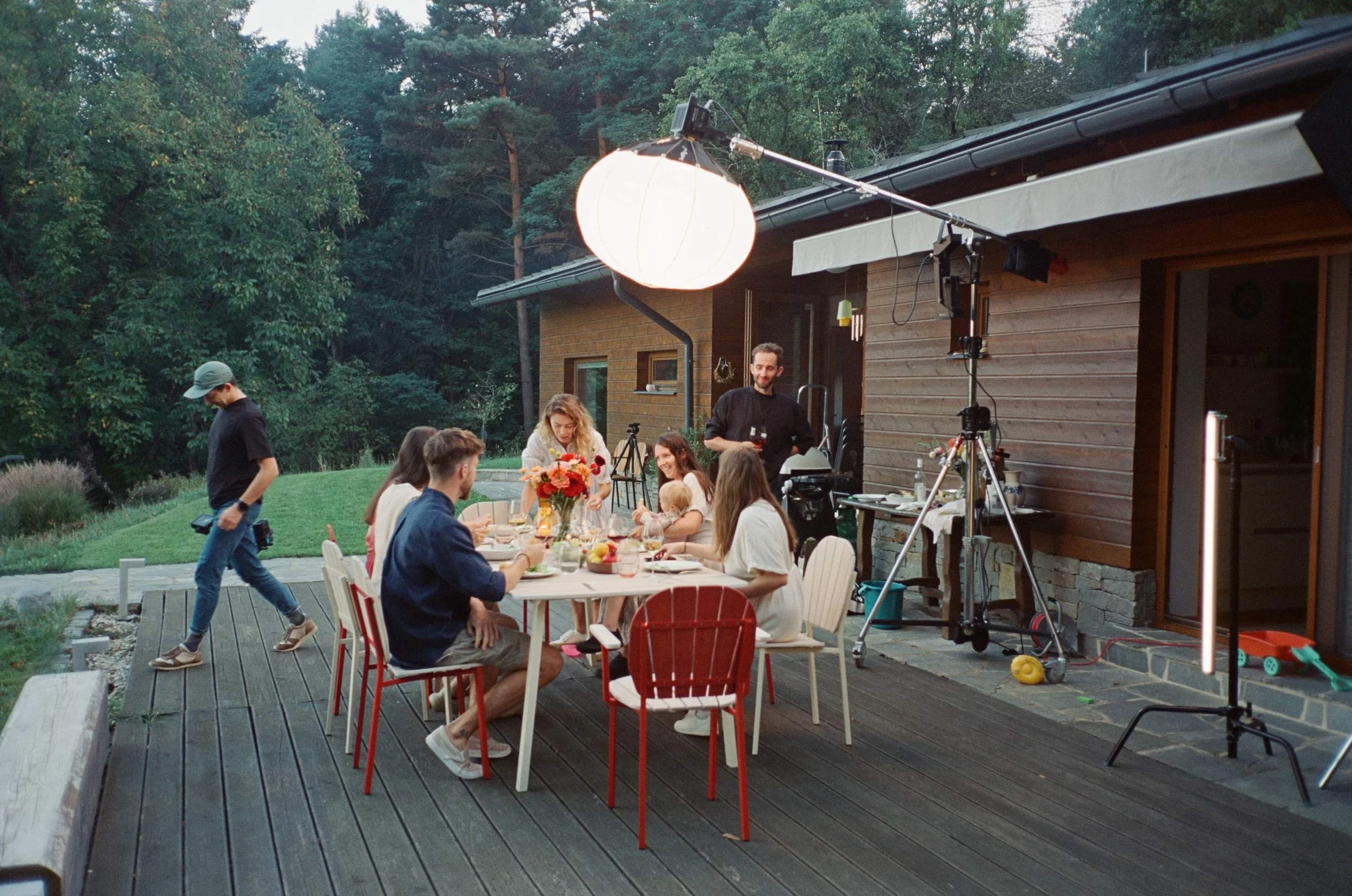 People gathered around a table for a dinner party outside at dusk, with professional lighting and film equipment set up for a shoot.