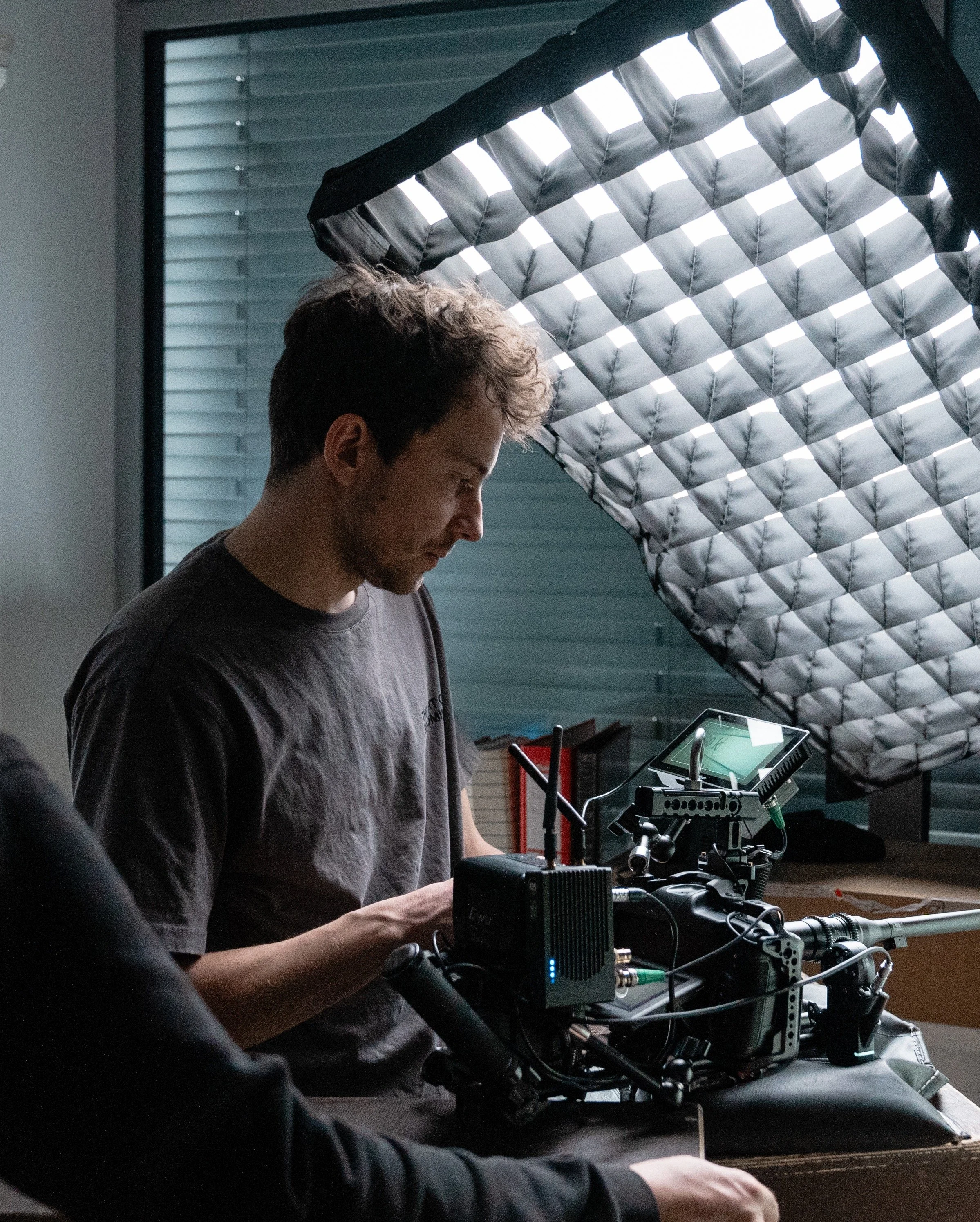 A person operating a professional camera setup in a dimly lit room, with a window and a partially opened window blind in the background.