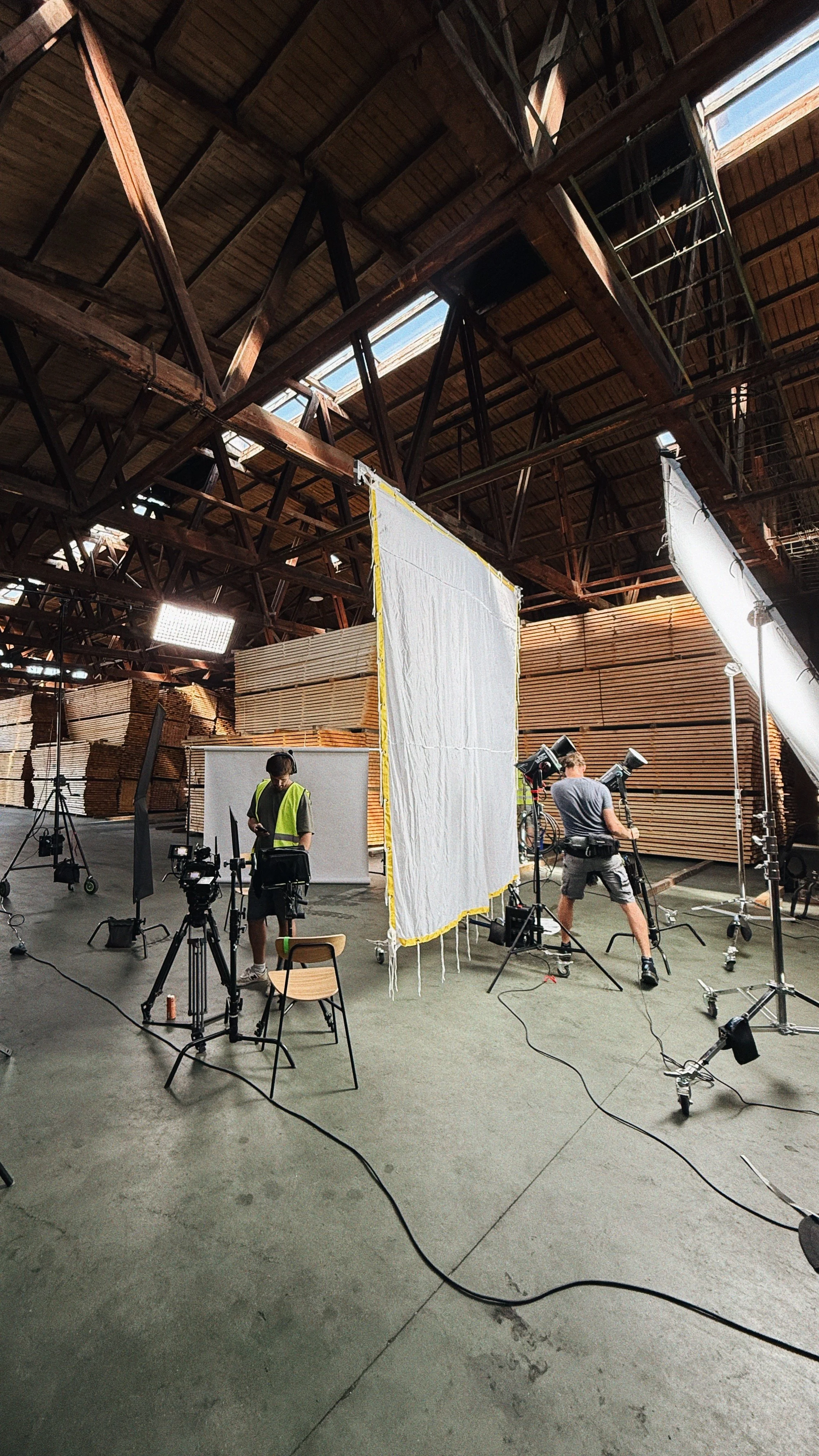 Film crew setting up equipment for a video shoot inside a lumber warehouse, with wooden planks stacked in the background.