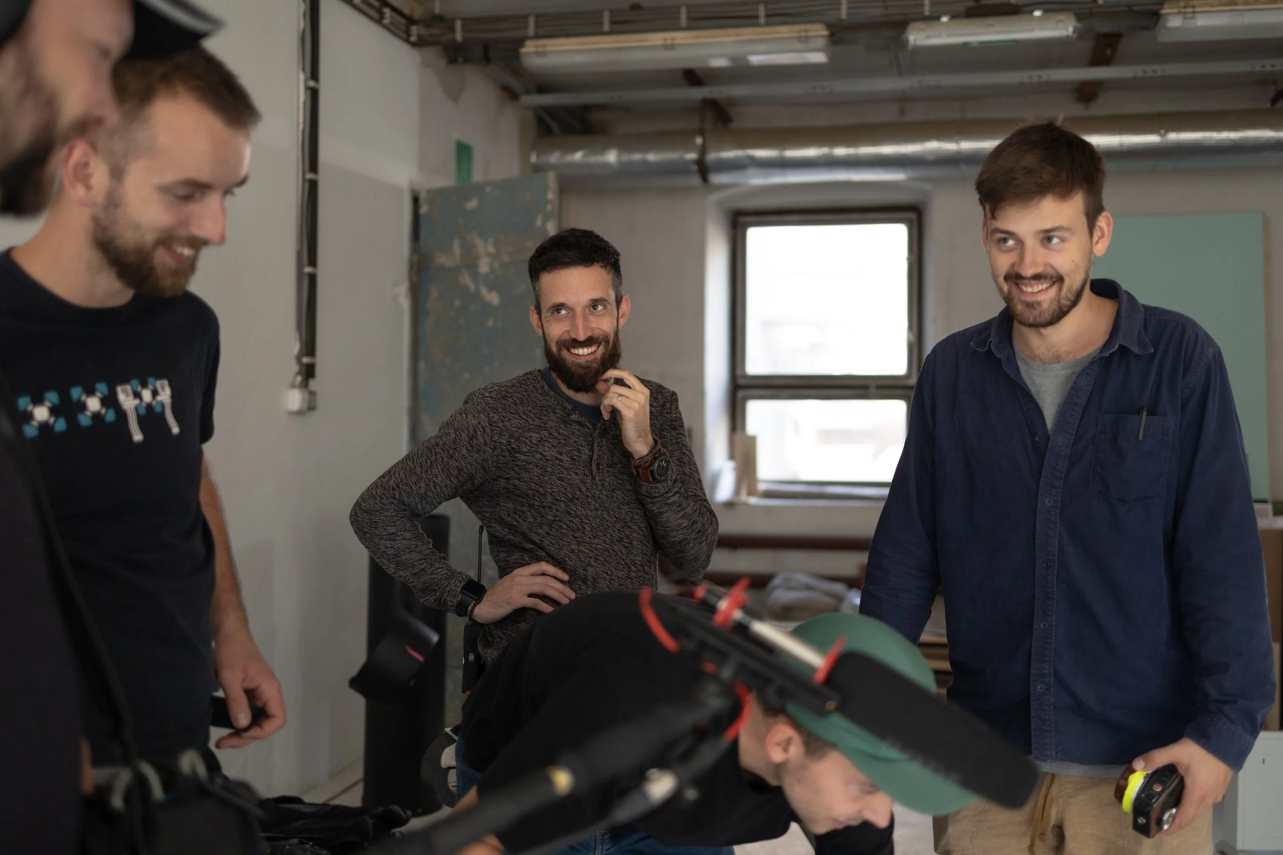 Four men in a well-lit room engaged in a conversation, one leaning over a piece of equipment while the others stand nearby smiling and talking.