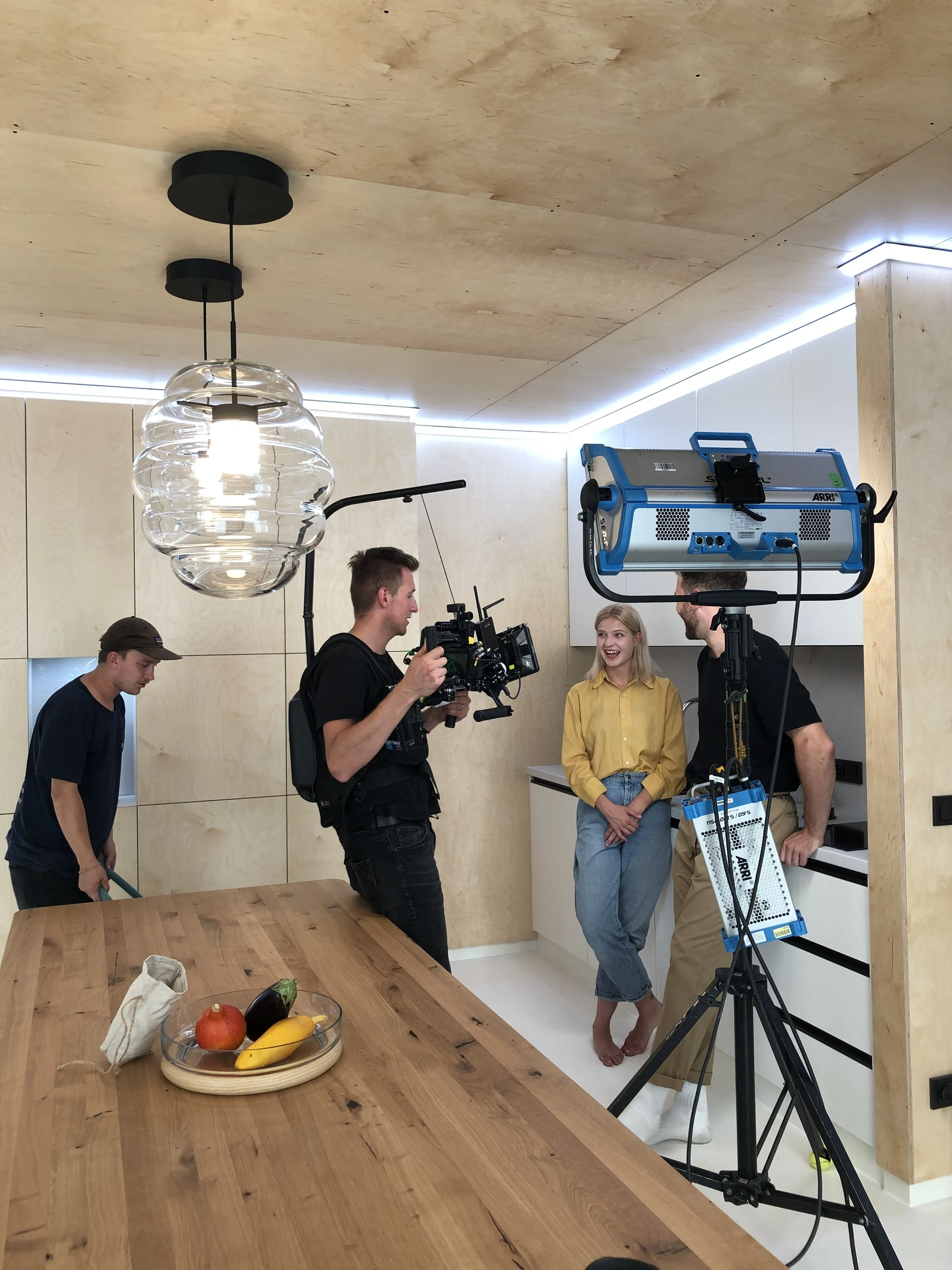 A film crew is filming a woman in a kitchen, with one crew member operating a camera and another adjusting lighting. A third crew member is cleaning the floor near the table with fruits.