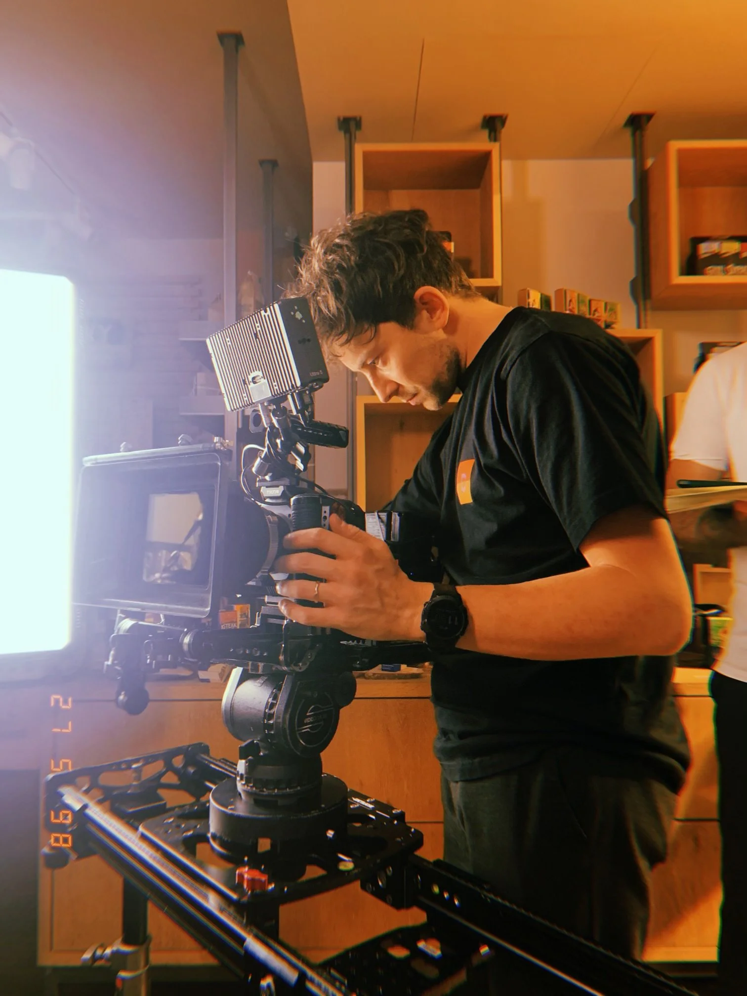 A man operating a professional camera rig in an indoor environment, with shelves and a bright light source in the background.