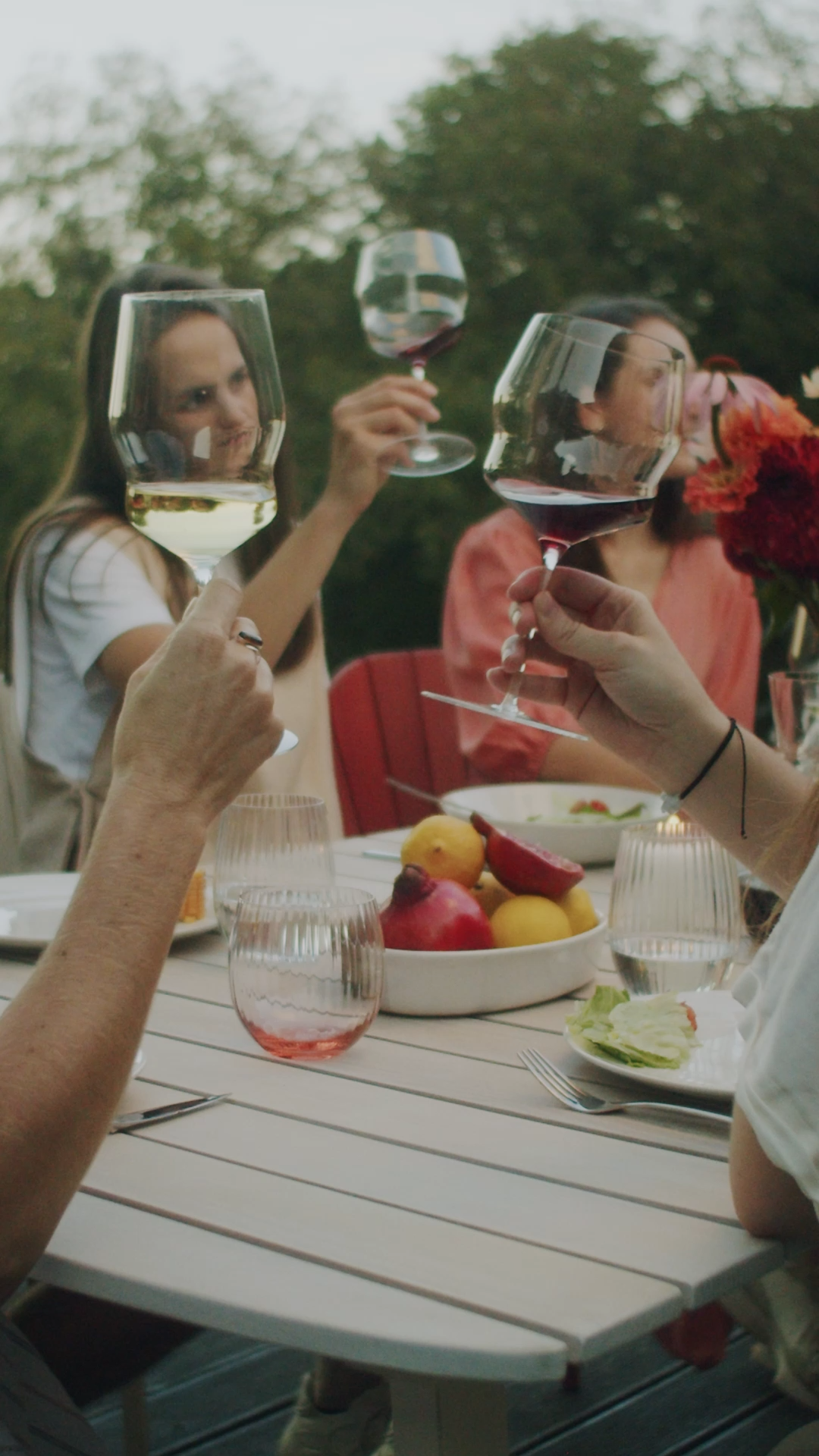 People clinking wine glasses at an outdoor dinner table with fruit, salad, and flowers.