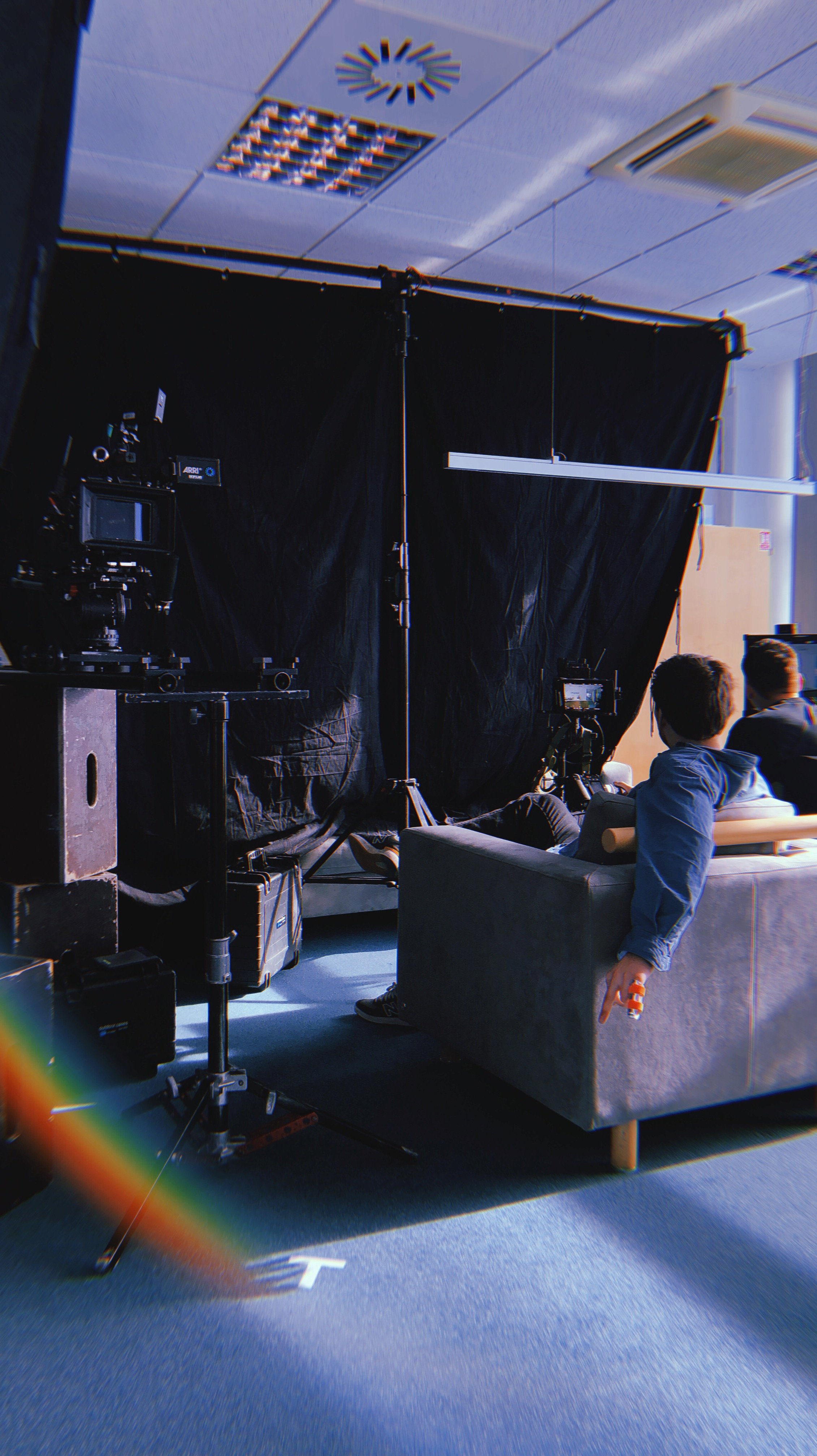 Indoor scene of a TV or film production set with cameras, lights, and a black backdrop, featuring two people seated on a couch, facing away from the camera.