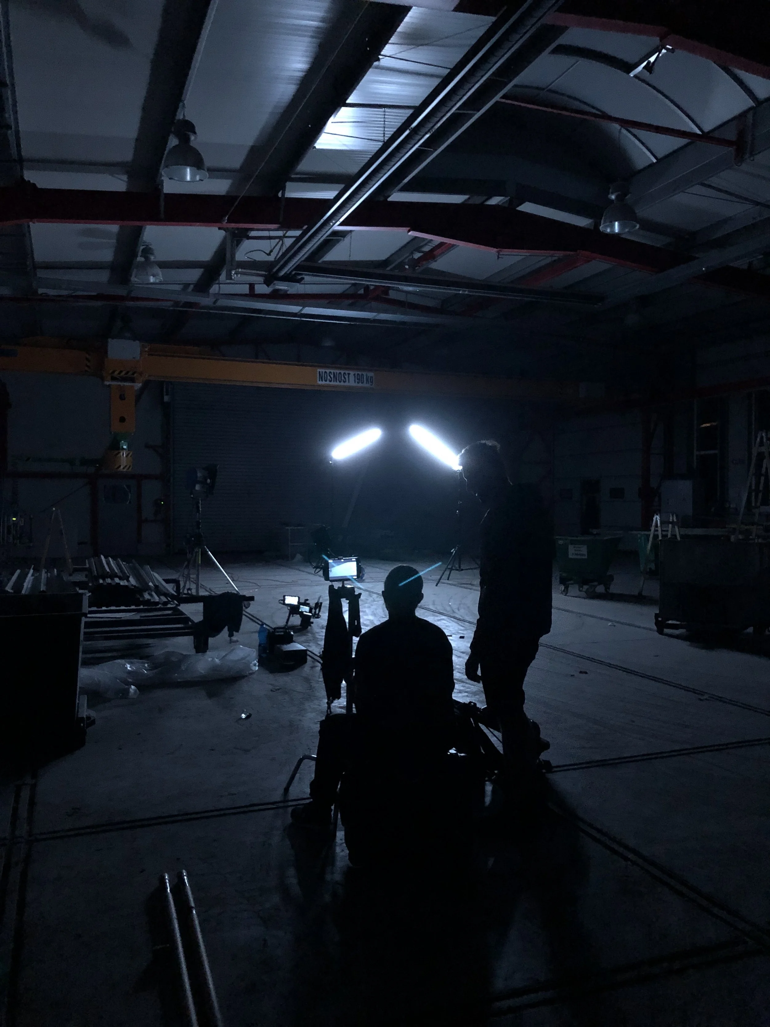 Film crew working in a dimly lit warehouse with equipment and a bright light source in the background.