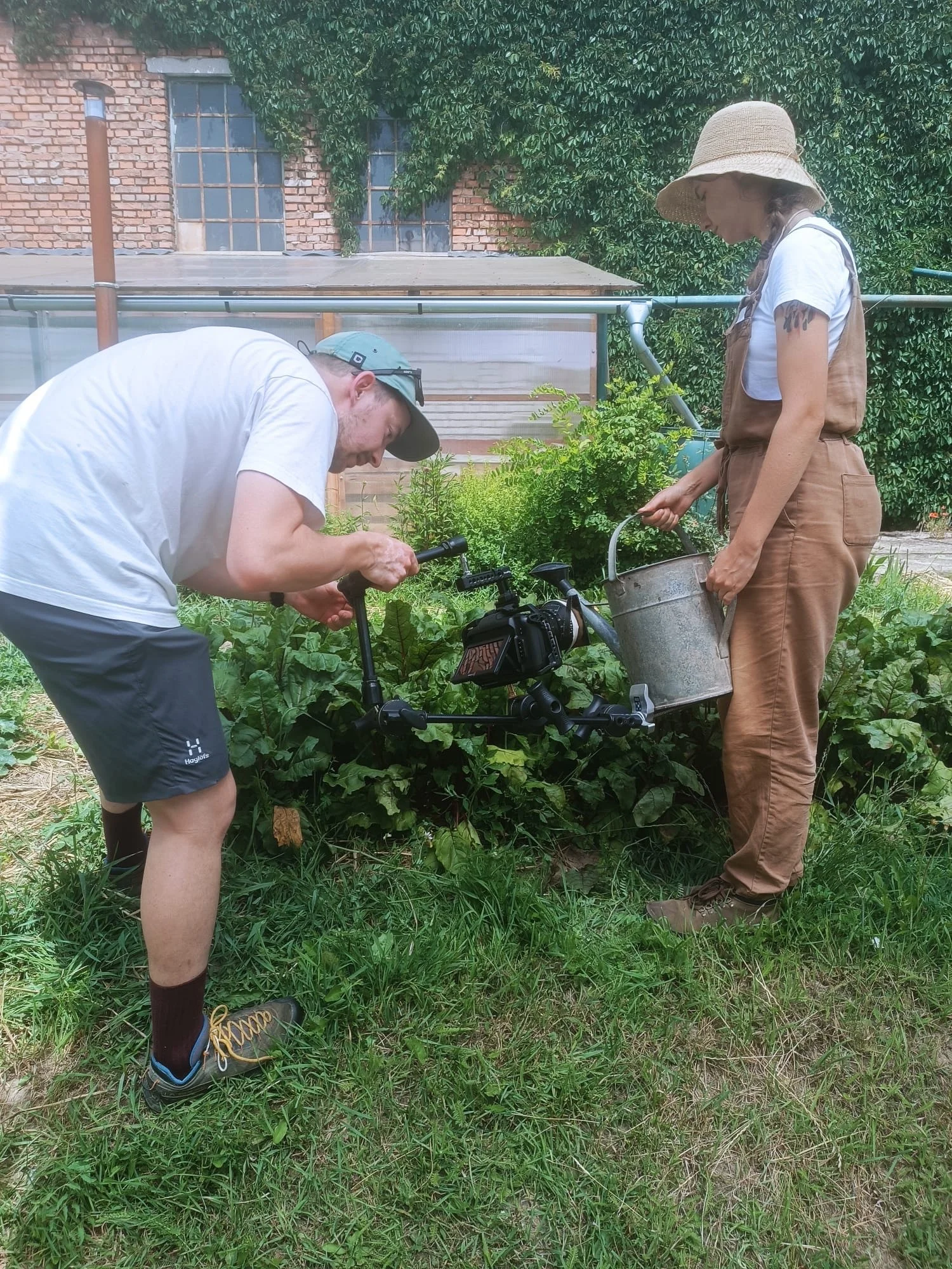 Two people working in a garden, one holding a watering can and the other adjusting a camera on a tripod.