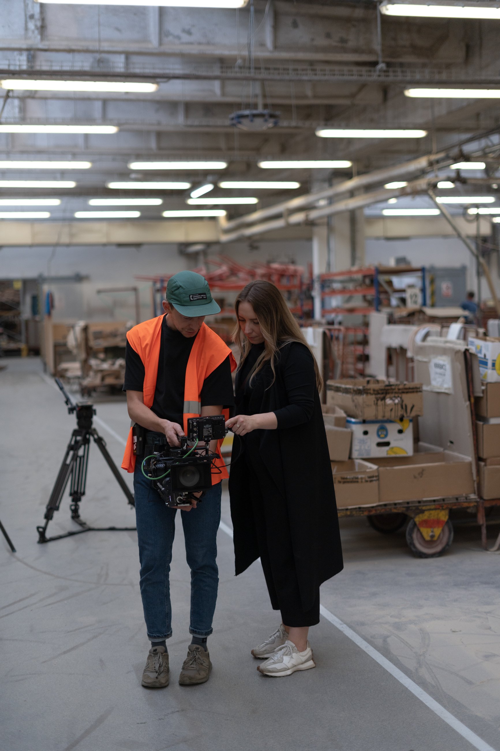 Two people looking at a camera inside a warehouse or factory with boxes and equipment around them.
