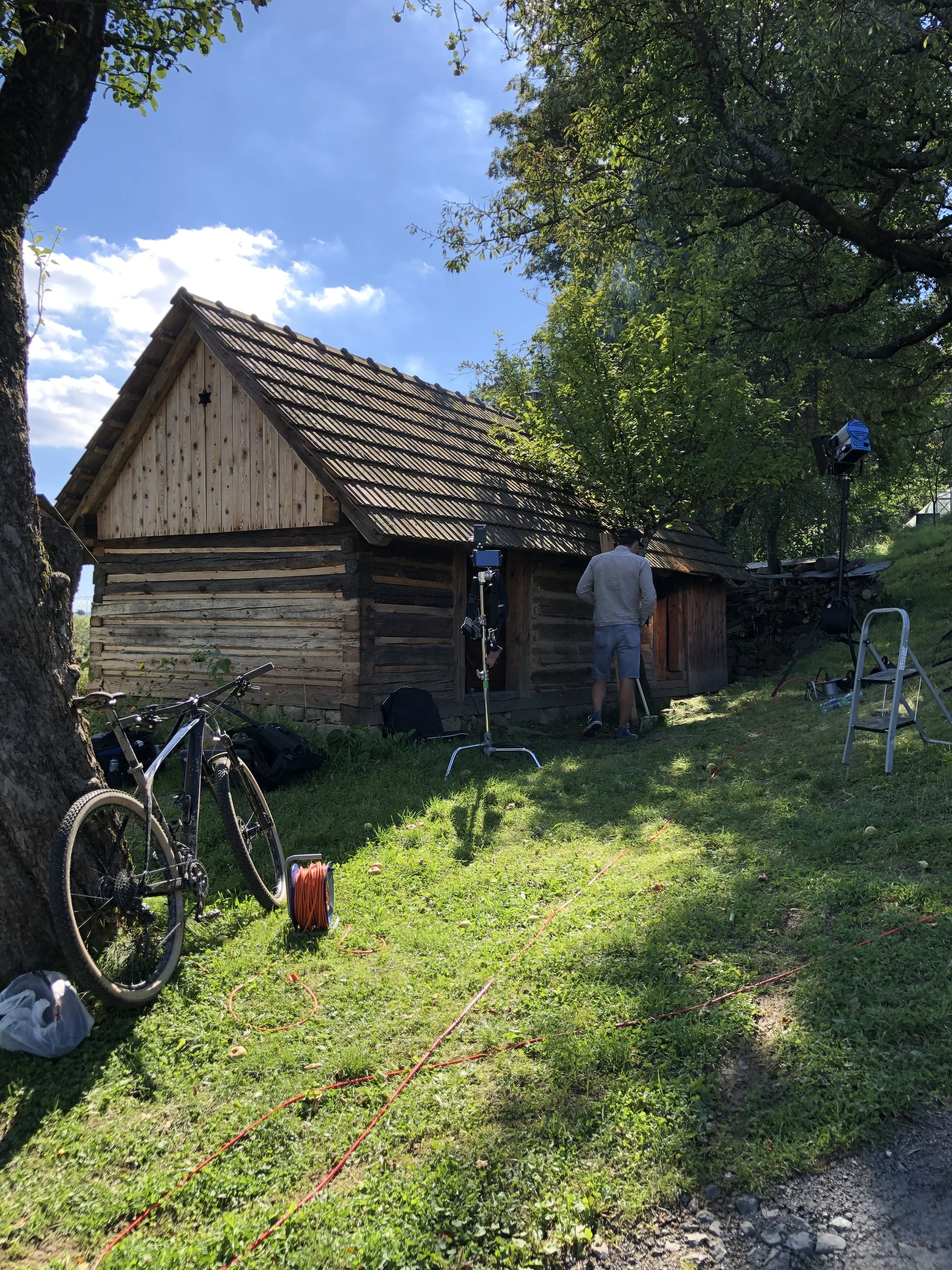A man working on a wooden cabin outdoors, surrounded by trees with a camera and lighting equipment set up around him, on a bright sunny day.
