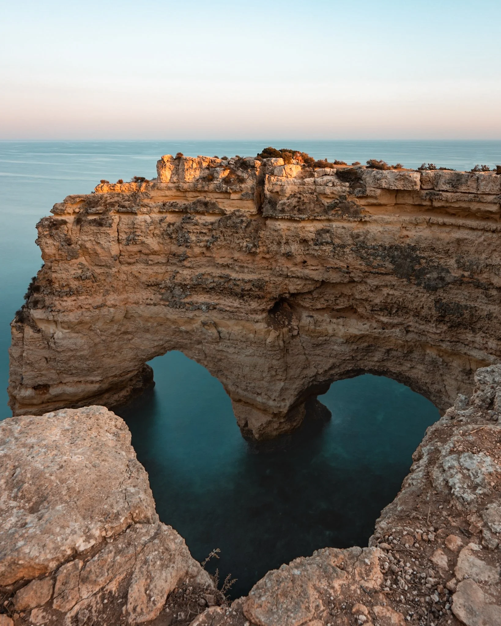A coastal cliff with two large natural arches over the ocean, with a clear sky in the background.