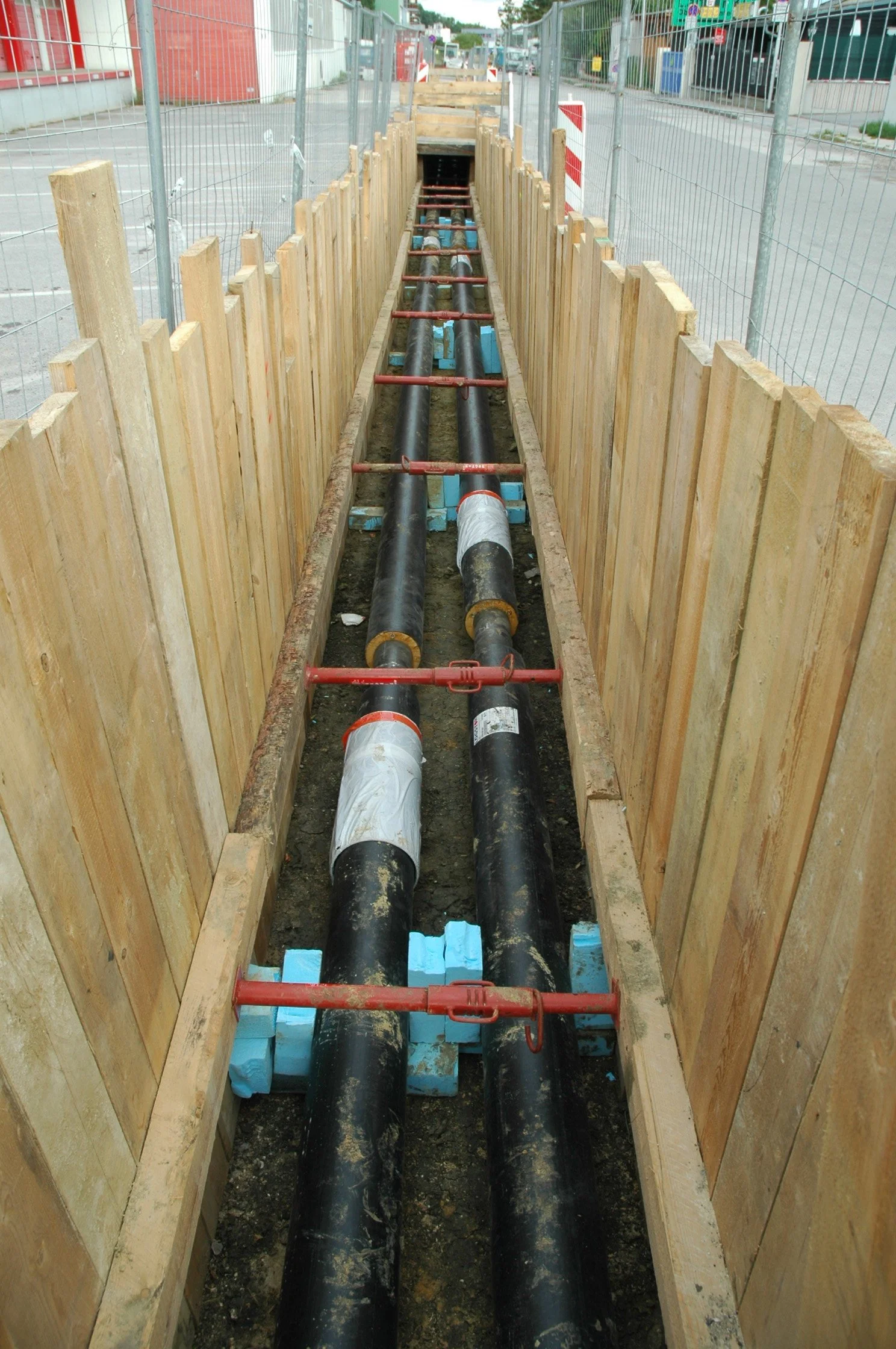 Underground piping installation at a construction site, with large black pipes supported by blue foam blocks, surrounded by wooden and metal safety barriers.