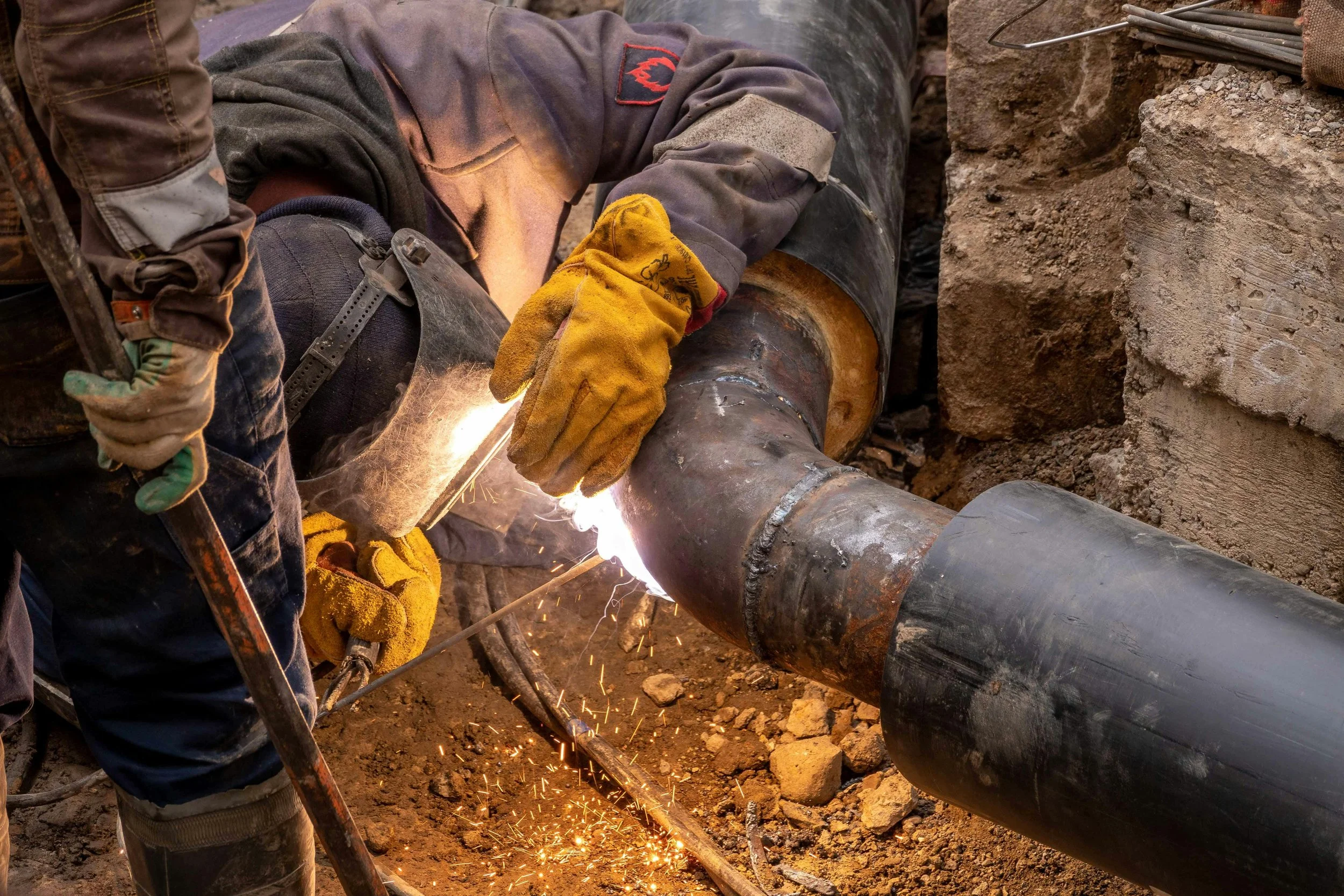 A worker wearing a helmet, gloves, and protective gear welding a large pipe underground, sparks flying during the process.