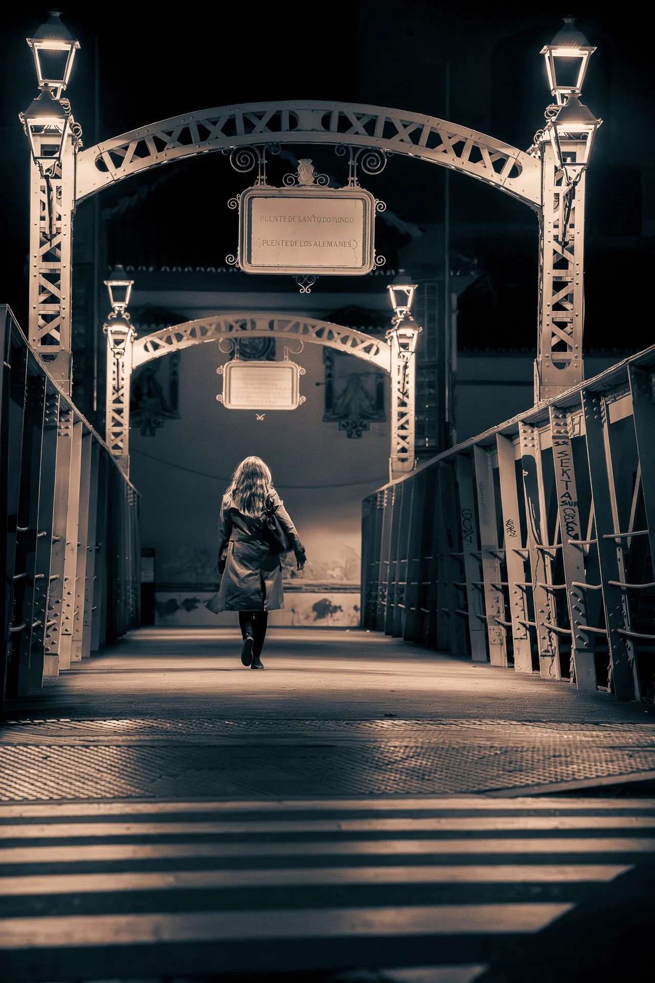 A woman walking alone on a bridge at night, illuminated by street lamps, with a sign overhead that reads 'Puente de Santo Domingo'.