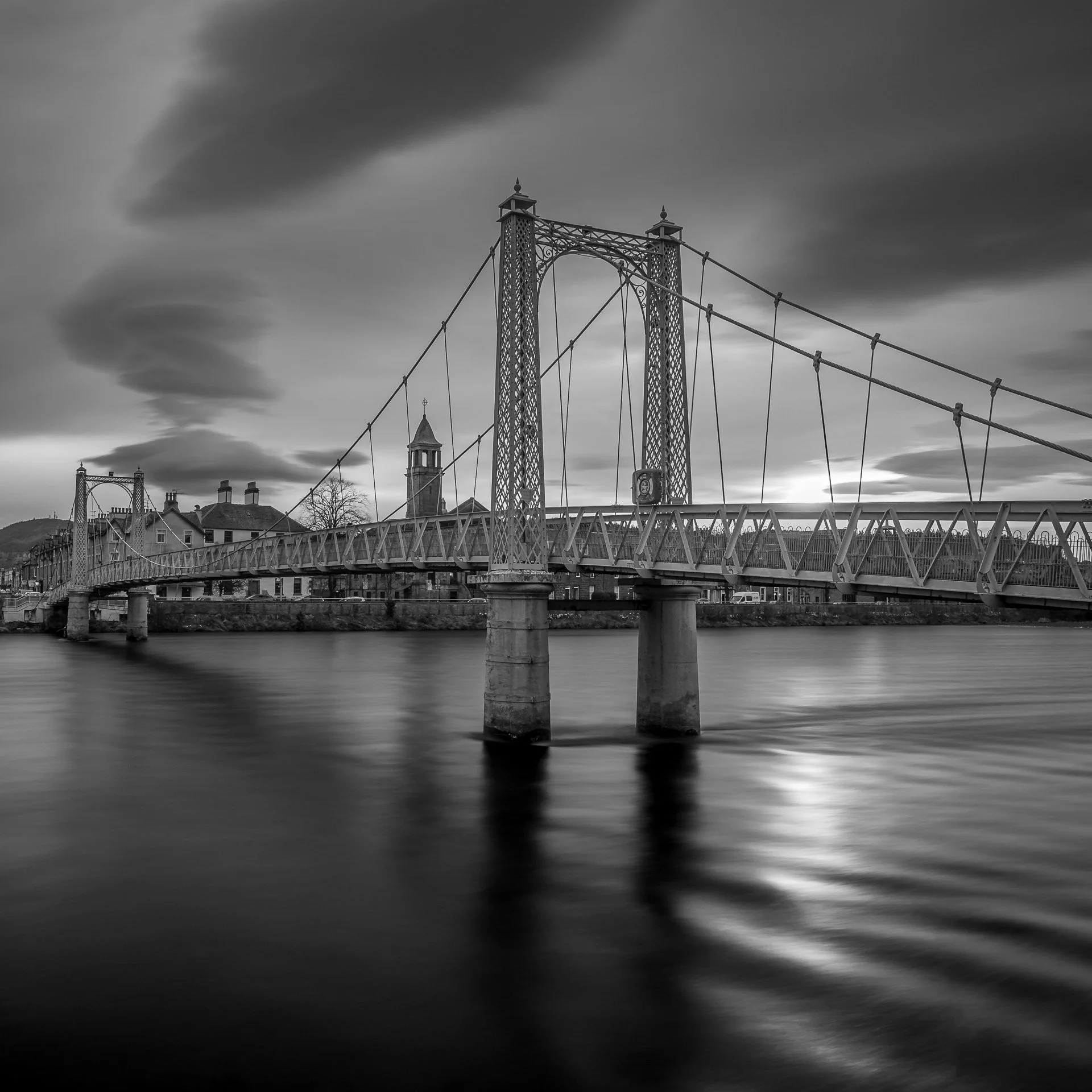 Black and white photo of a suspension bridge over a river with buildings and trees in the background, under a cloudy sky.