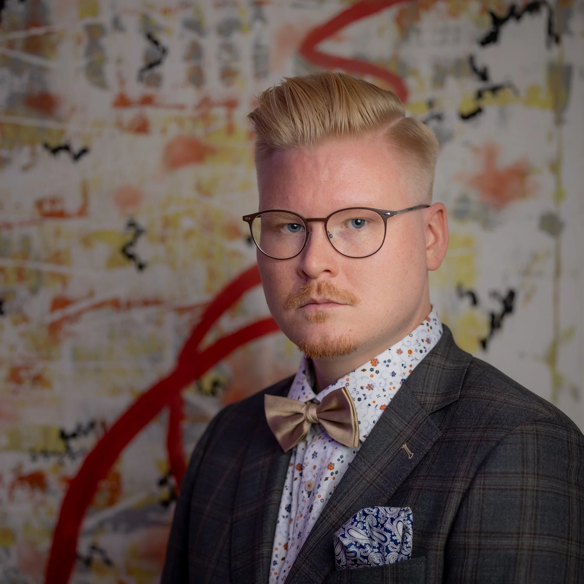 Young man with short blonde hair, glasses, wearing a dark plaid suit, patterned dress shirt, and beige bow tie, standing in front of graffiti-covered wall.