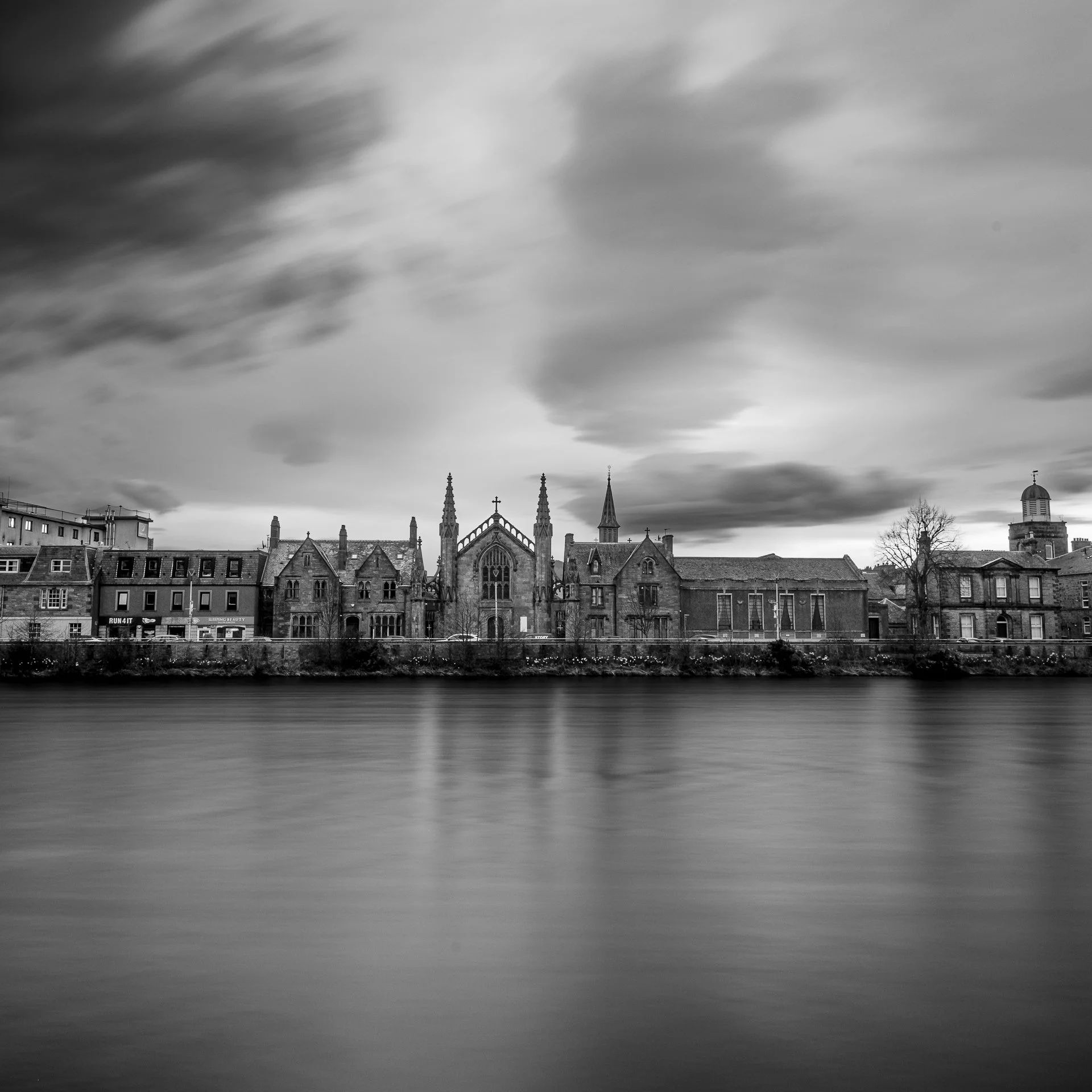 Black and white photo of a cityscape with historic buildings and church spires under a cloudy sky, reflecting on a body of water.