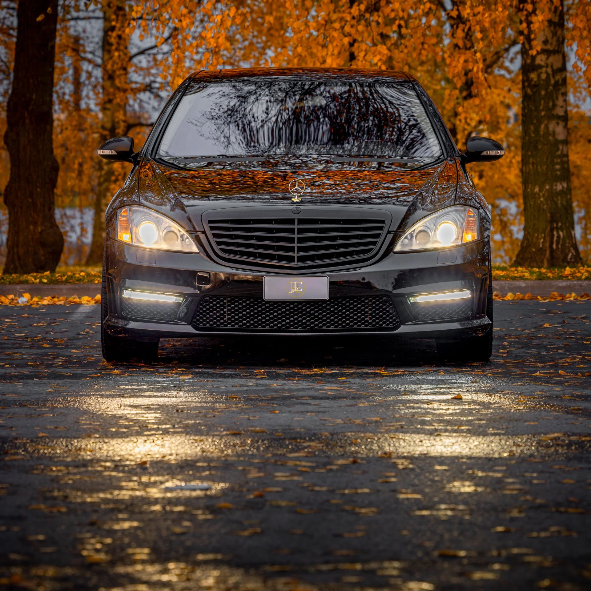 A black Mercedes-Benz car parked on a paved road amidst fallen autumn leaves, with a background of trees with orange and yellow foliage, reflected on the car's surface.