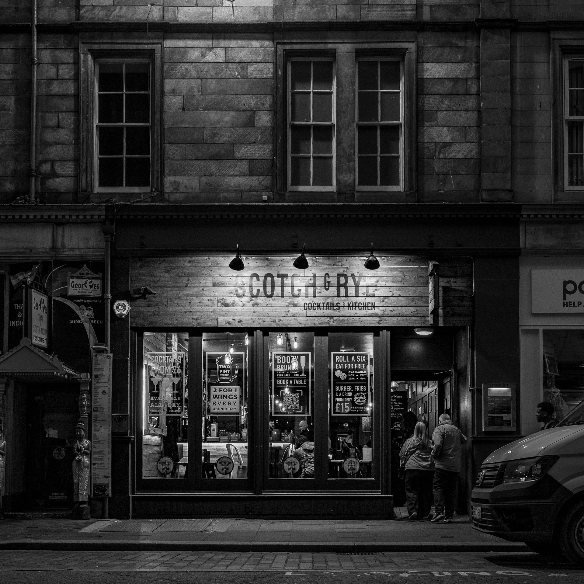 Black and white photo of a street view showcasing a two-story building with large windows and a sign that reads 'Scott & Gory Cocktails & Kitchen.' Several people are walking and standing near the entrance, and there are various signs, lights, and objects outside the establishment.