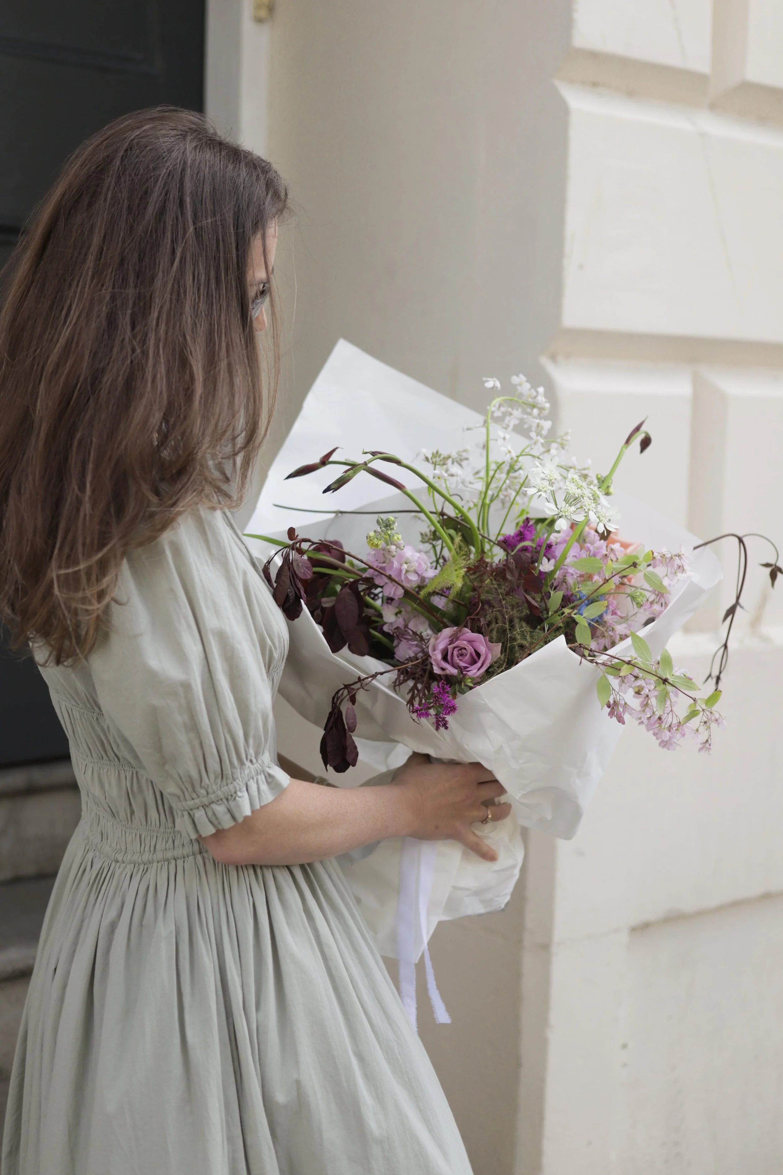 A woman with long brown hair wearing a light-colored dress holding a graduation bouquet of mixed flowers, including purple roses and white blooms, wrapped in white paper.