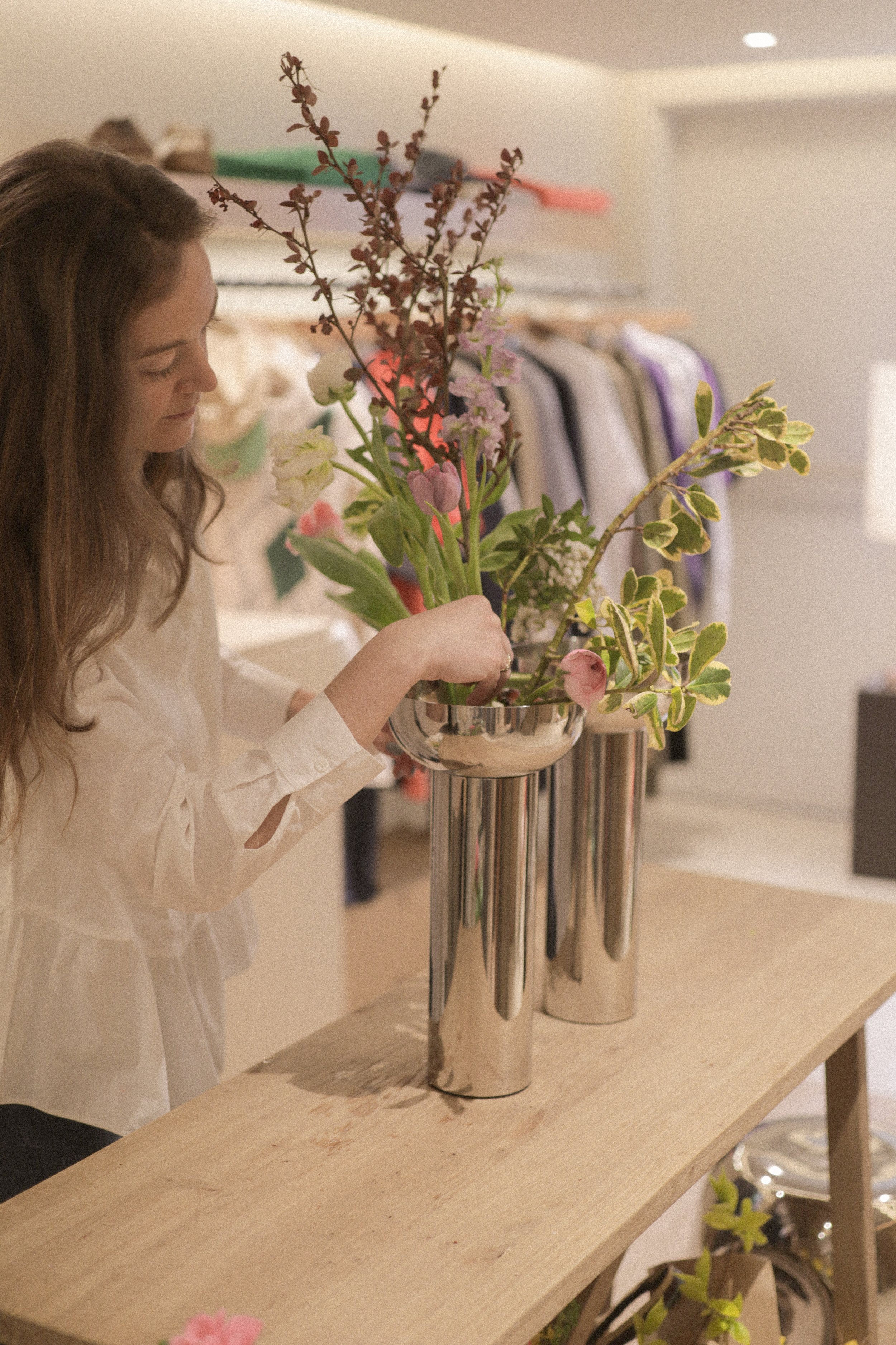 Woman arranging flowers in a metallic vase displayed on a wooden table. The background shows a clothing rack with various garments.