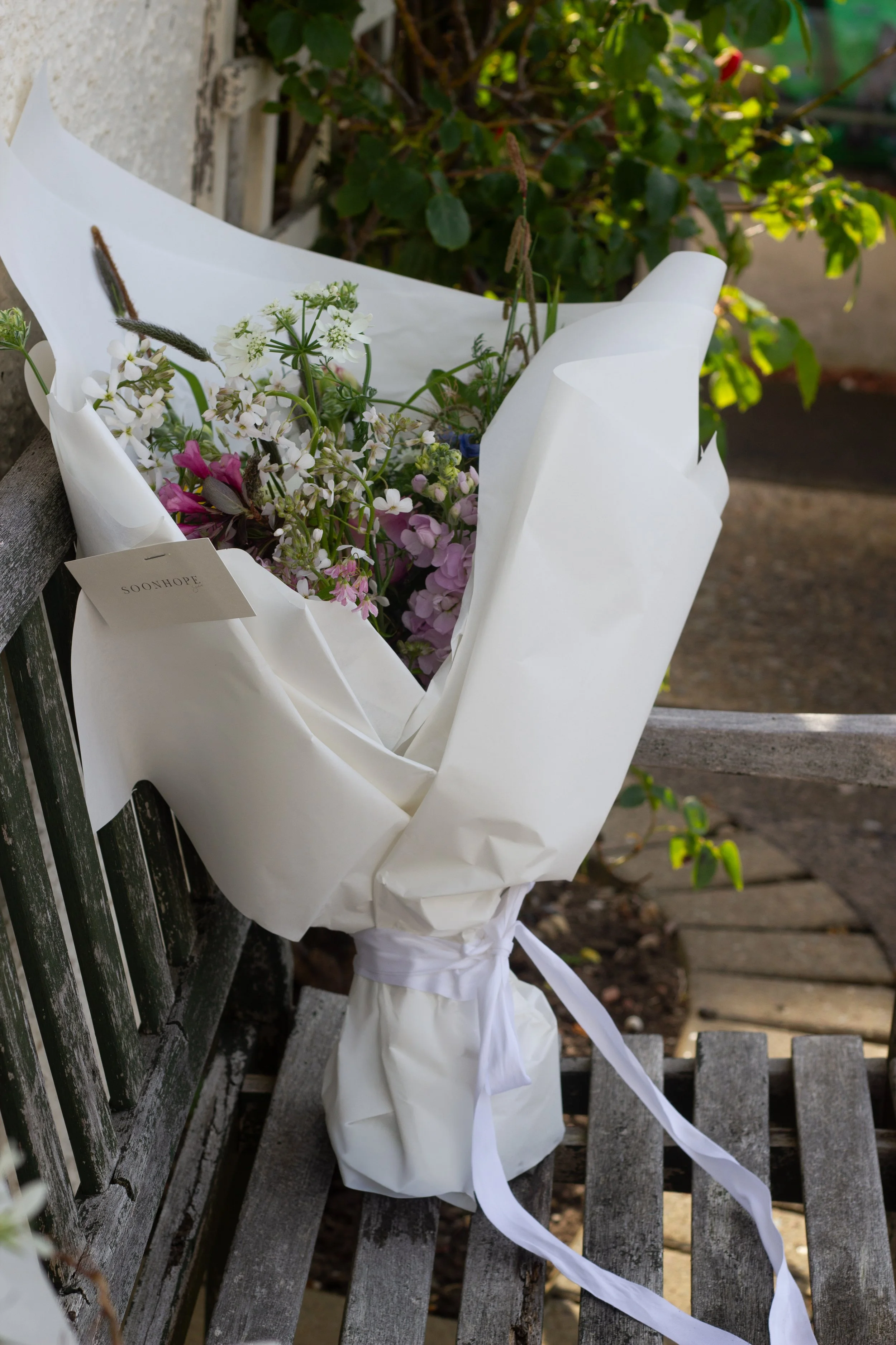 A bouquet of flowers wrapped in white paper, tied with a white ribbon, sitting on a weathered wooden bench outdoors.