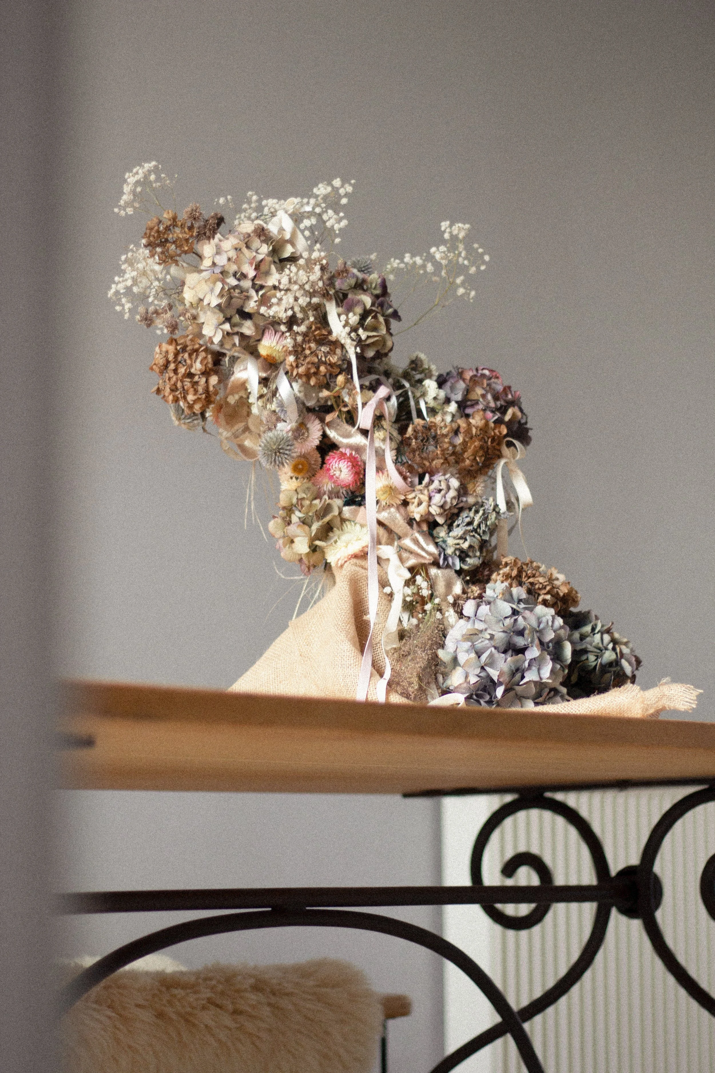 A dried flower arrangement with various flowers and ribbons, placed on a light-colored cloth, on a wooden surface against a gray wall.