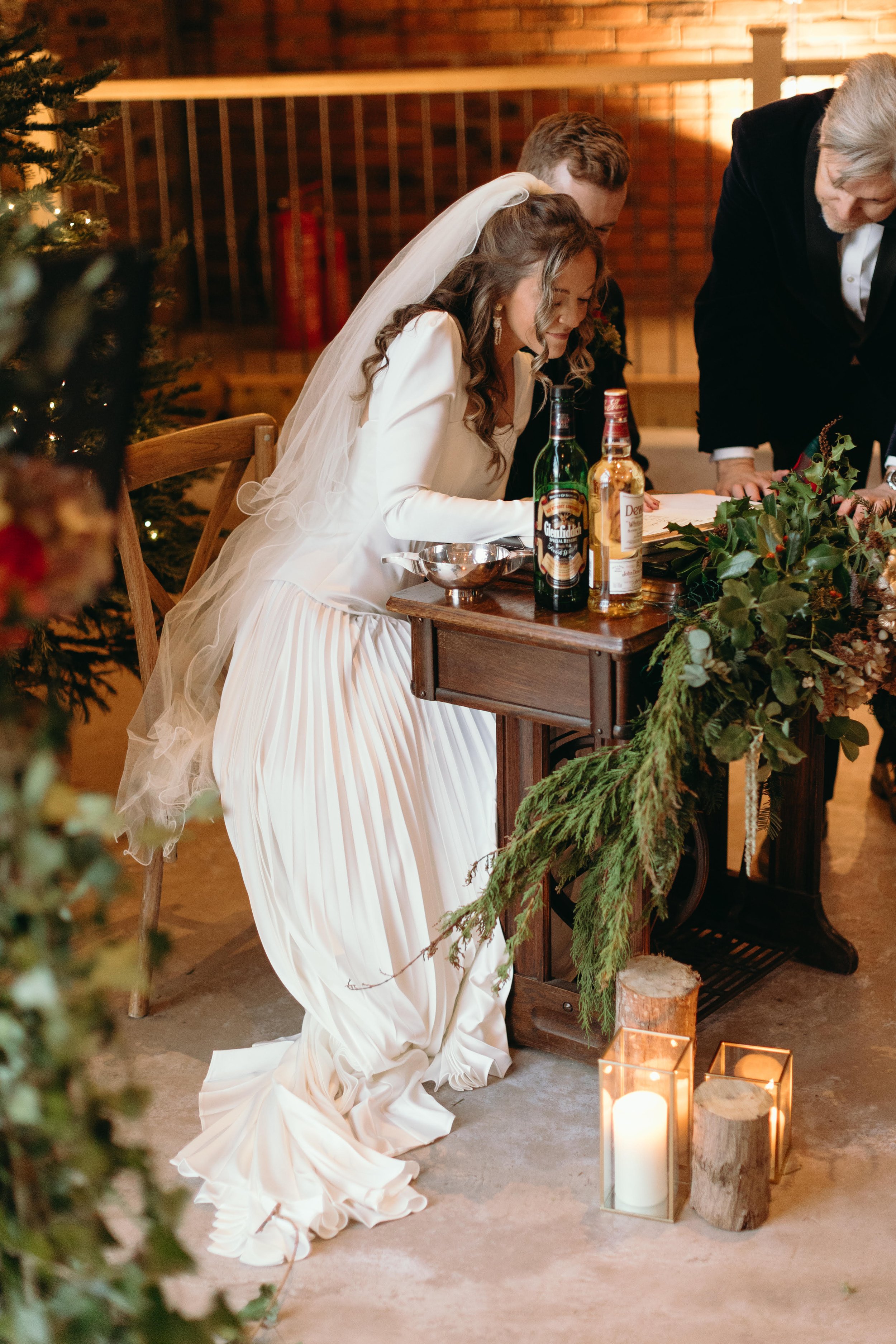 A bride in a white dress with a veil signs a document at a table decorated with greenery and candles, accompanied by two men in formal attire, in a rustic indoor setting with a brick wall background.