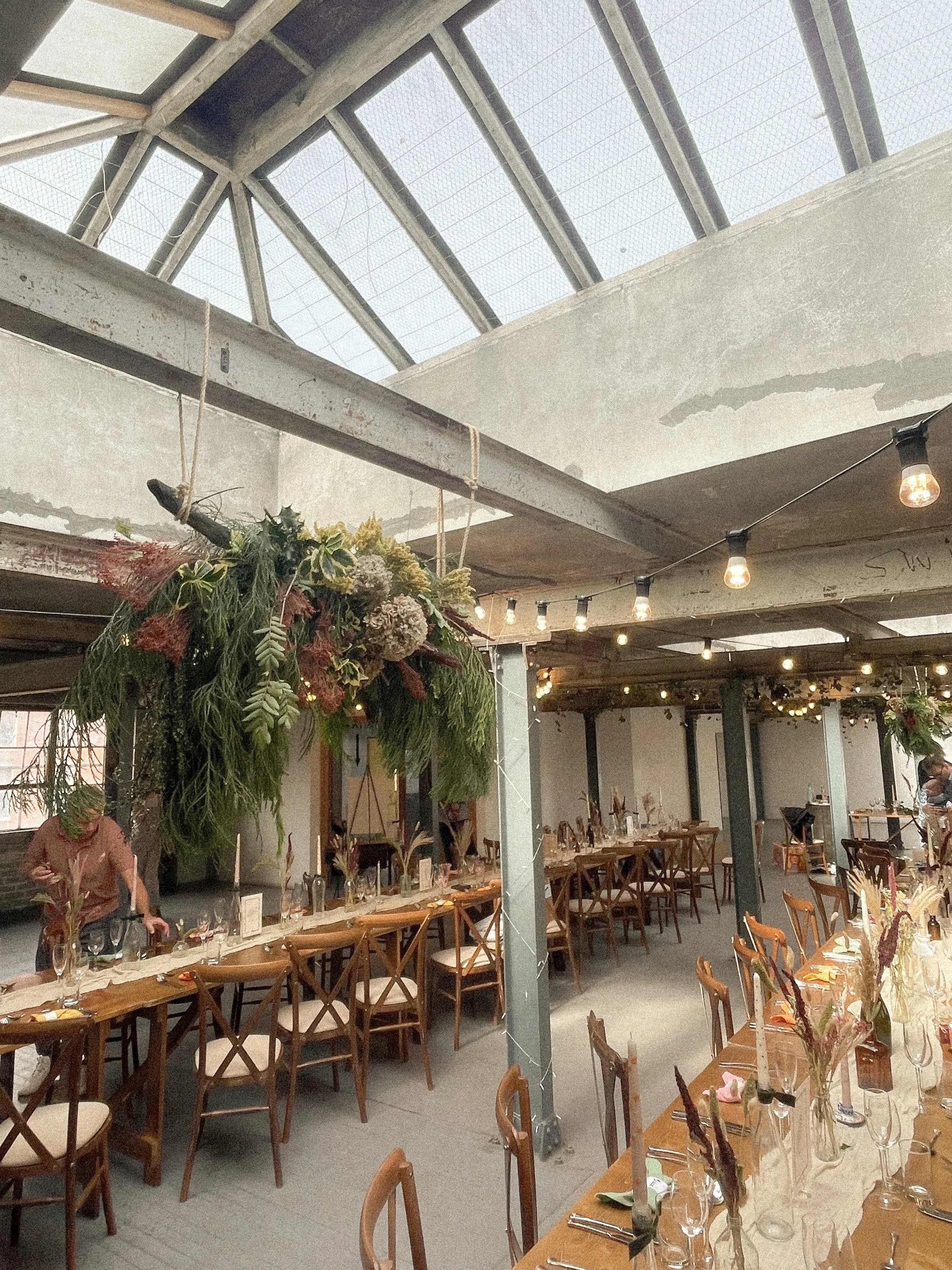 A decorated indoor wedding venue set for a meal with tables, chairs, floral centerpieces and hanging installations, string lights, and featuring a large windowed ceiling.