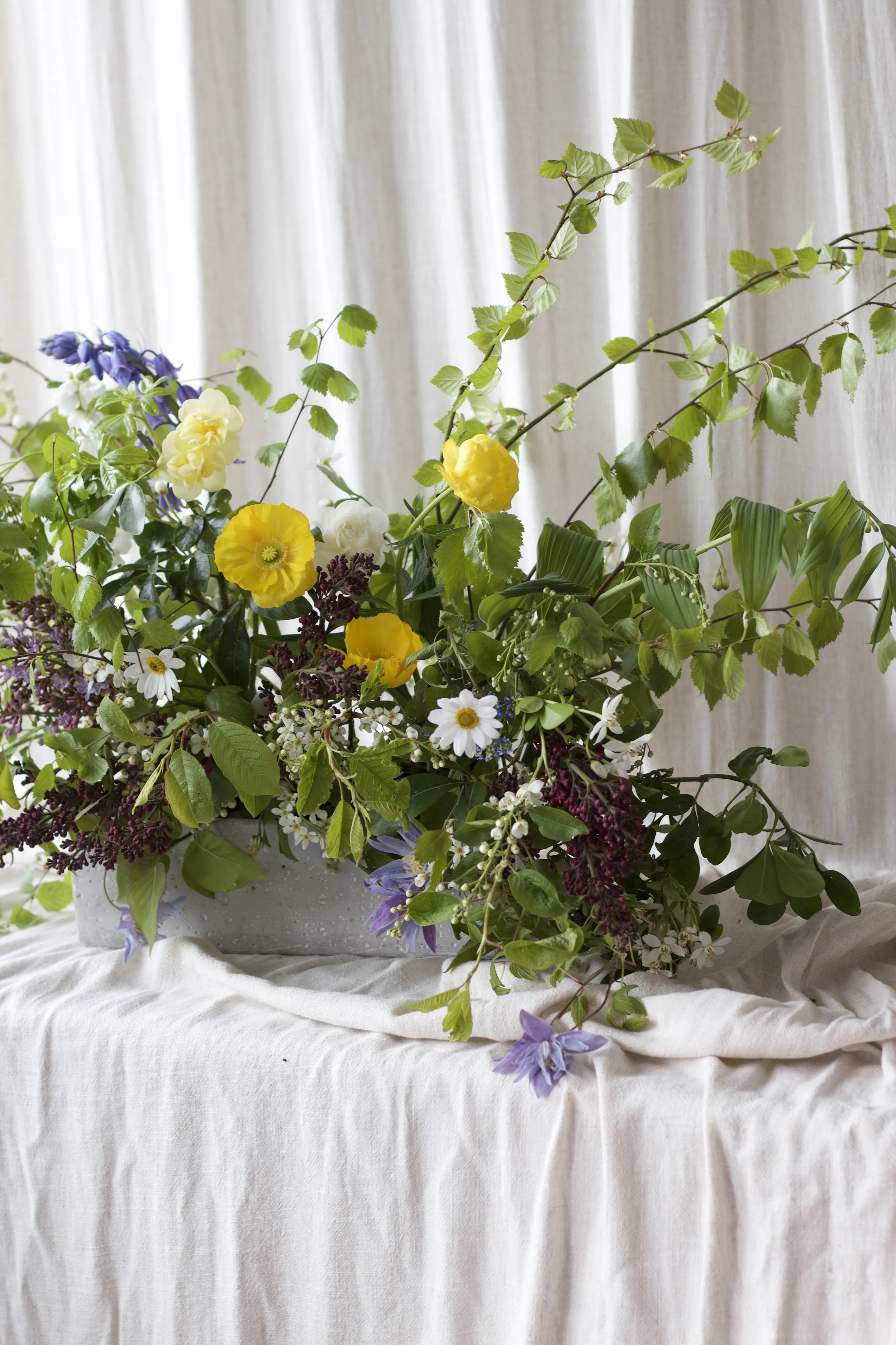A large floral arrangement, a ground meadow, with yellow, white, purple, and blue flowers and green leaves, displayed on a white tablecloth with a veiled background.