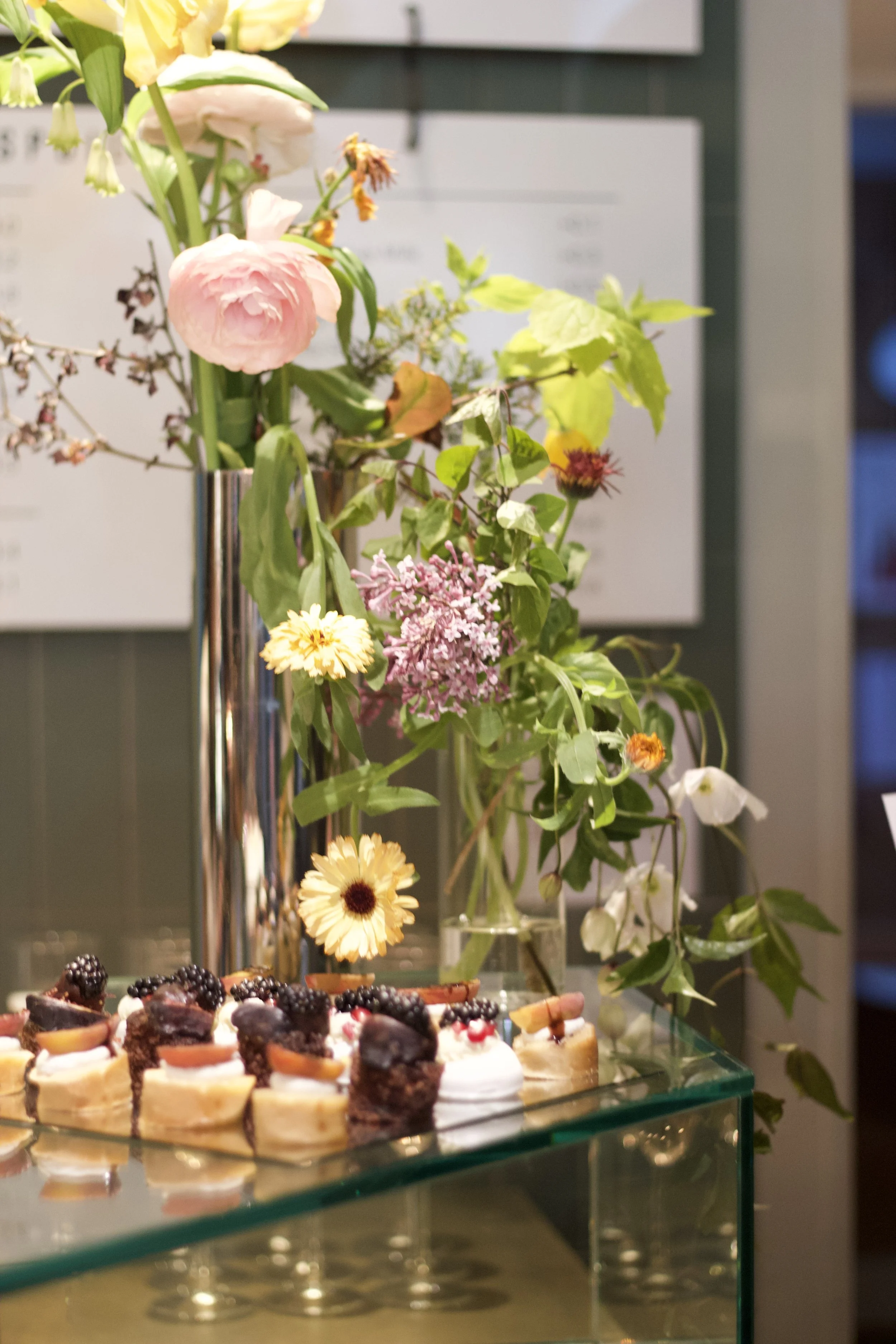 A floral arrangement with pink, white, and yellow flowers in a tall silver vase, placed behind a tray of assorted desserts on a glass table.