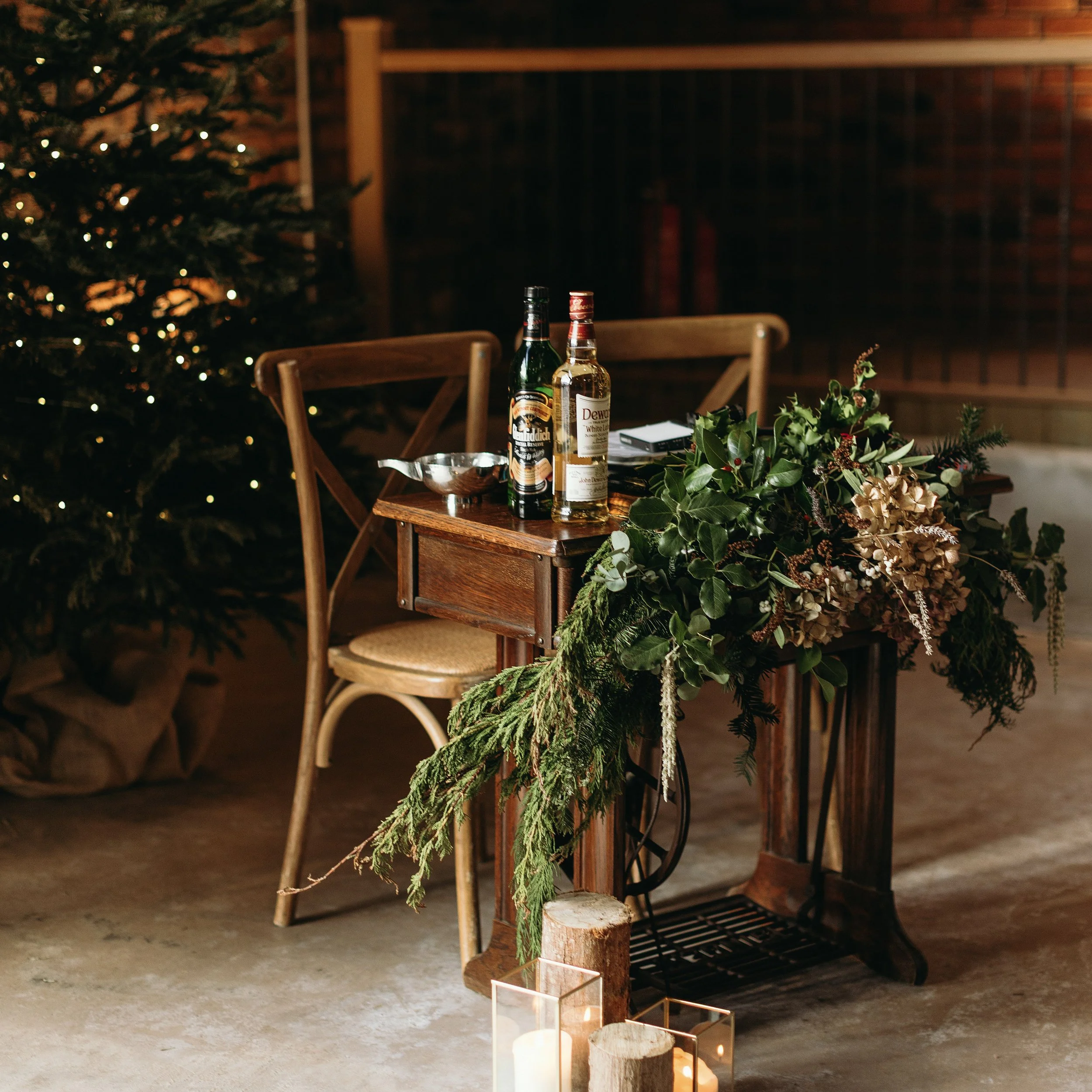 A decorated Christmas tree with lights in the background, and a wooden dining table adorned with Christmas greenery, candles, and bottles of alcohol.