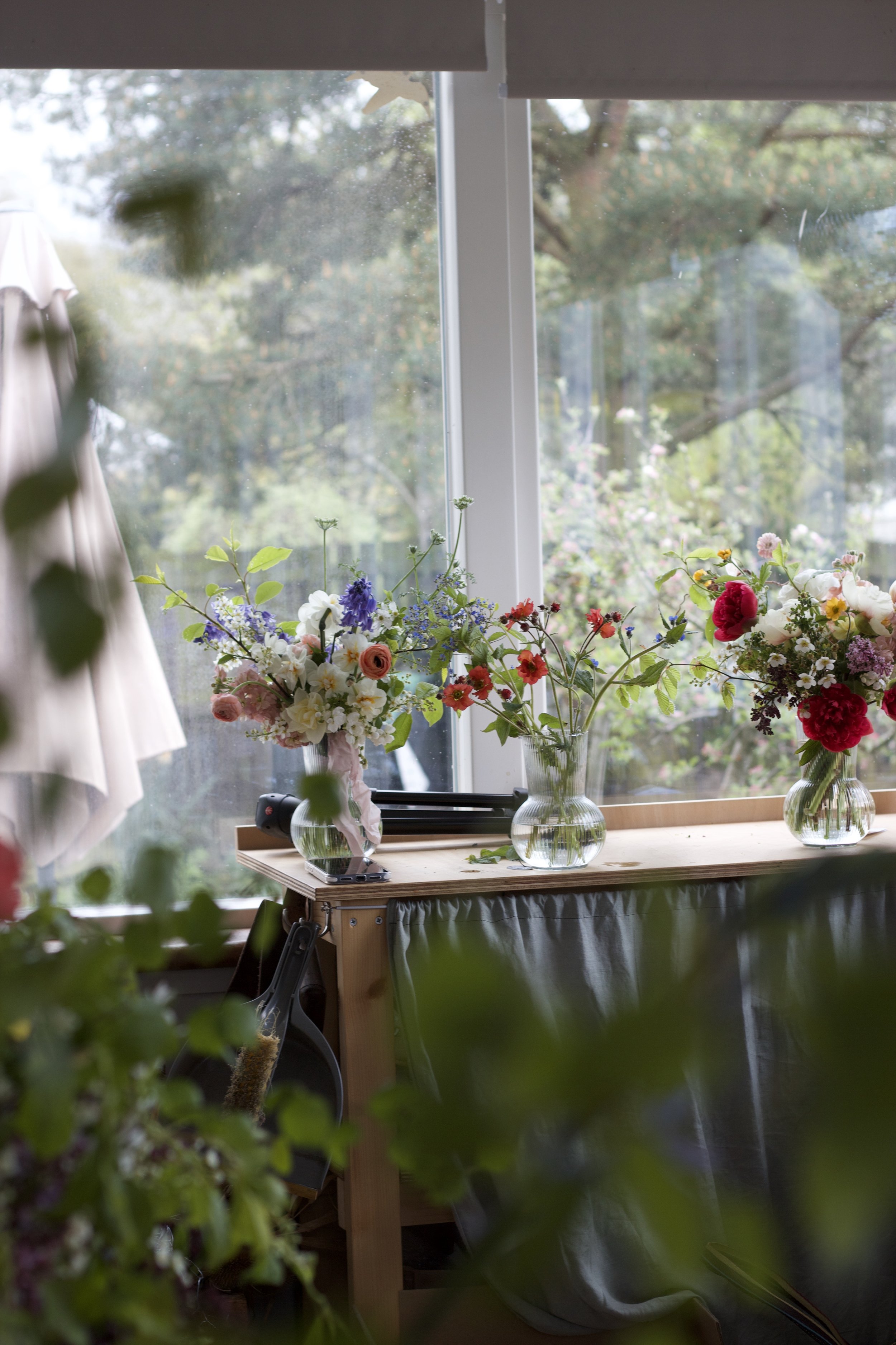 A windowsill with three glass vases filled with colorful flowers, with trees and a cloudy sky visible outside.