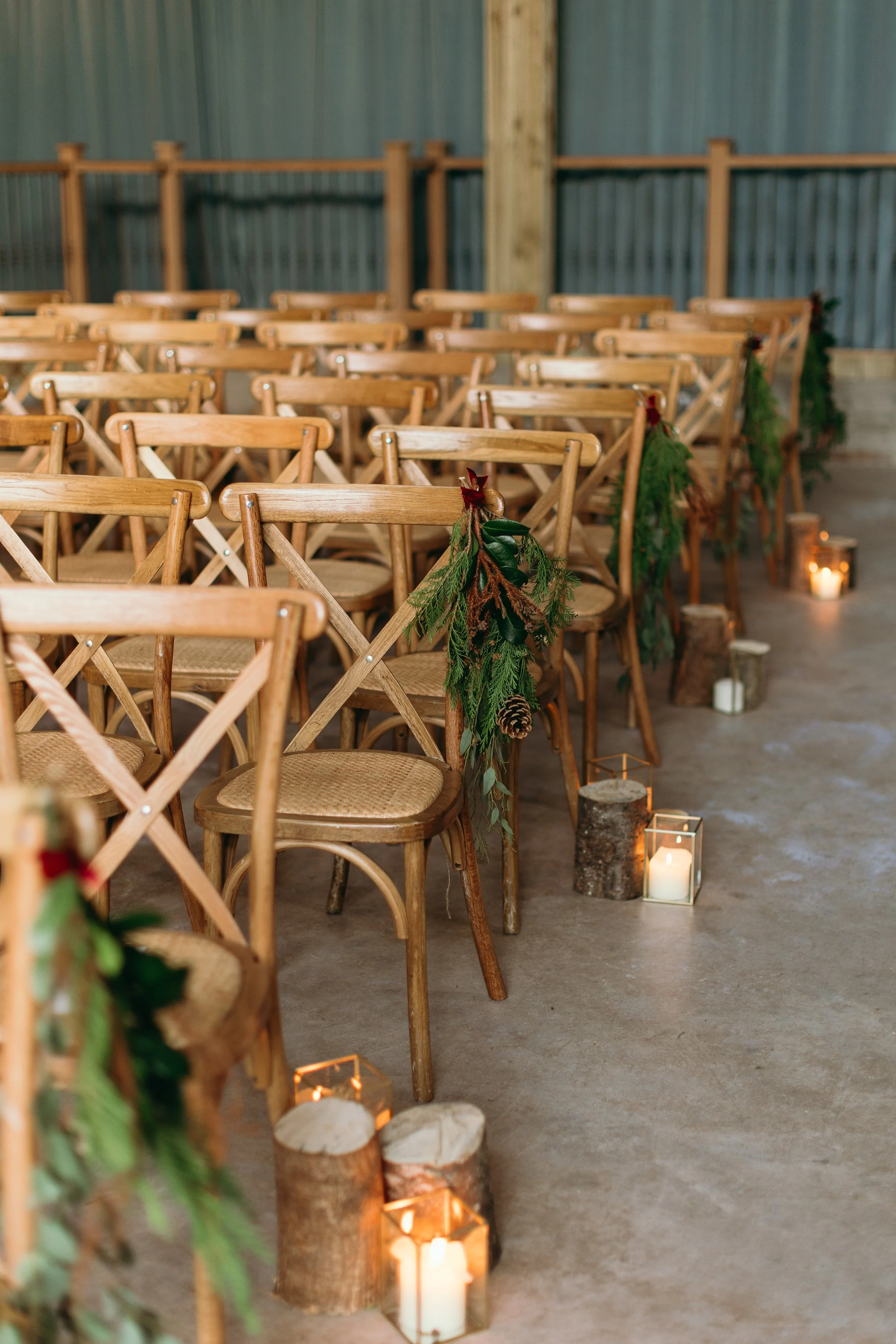 Wooden chairs decorated with greenery and pinecones, arranged for a gathering with lit candles in lanterns along the aisle.