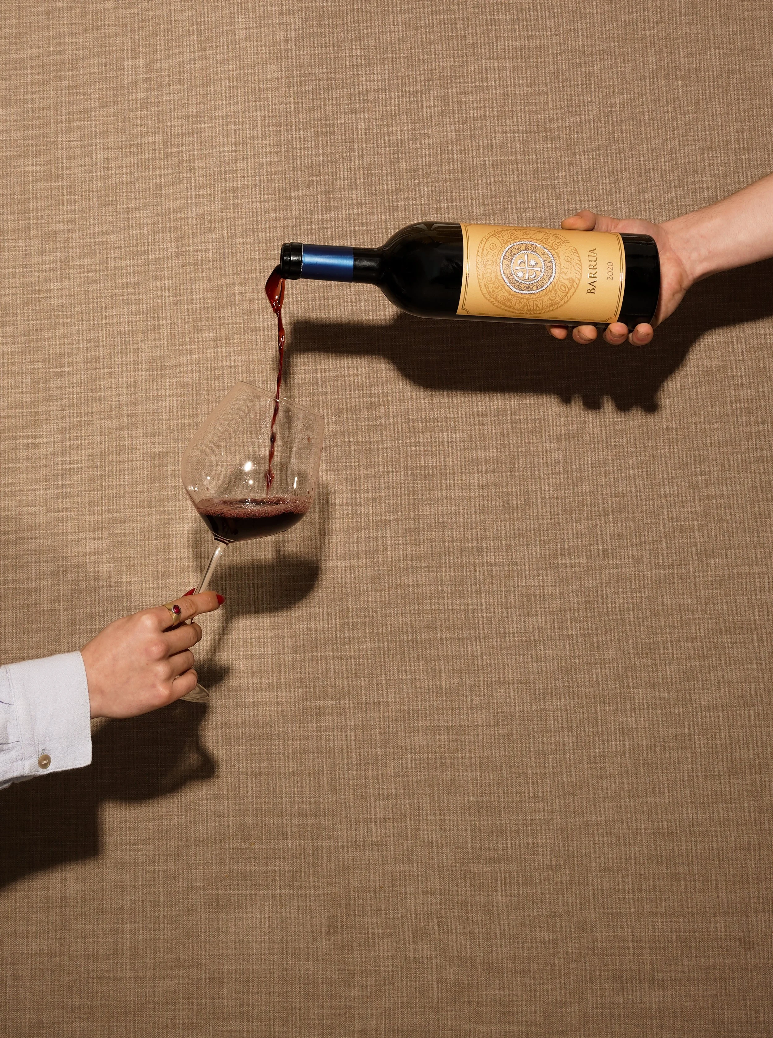 Main tenant un verre de vin rouge à une main, un autre main verse du vin rouge d'une bouteille dans le verre, fond beige