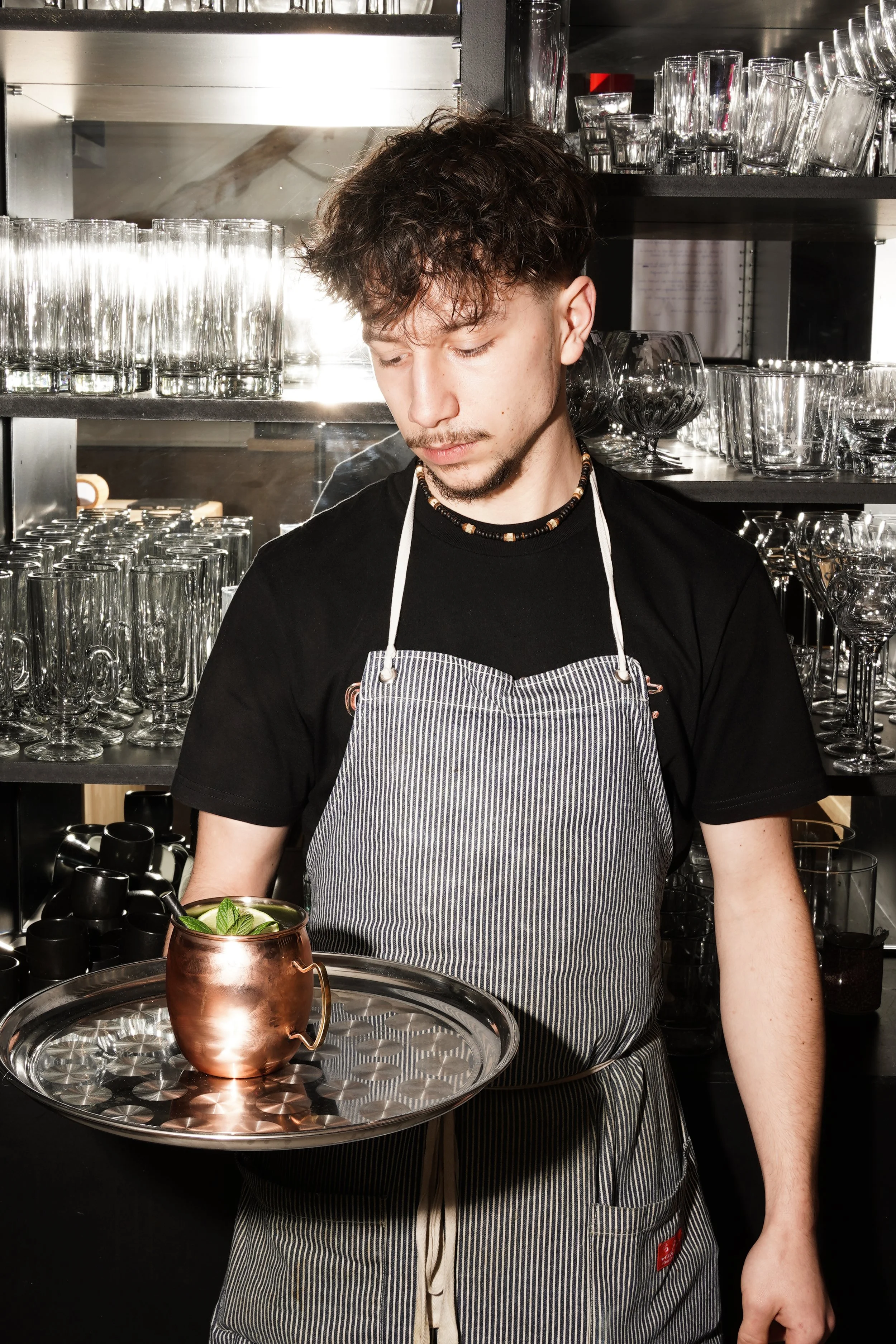 Jeune homme portant un tablier à rayures, tenant un plateau avec une tasse en cuivre contenant des glaçons et de la menthe, dans un bar ou un restaurant en arrière-plan.