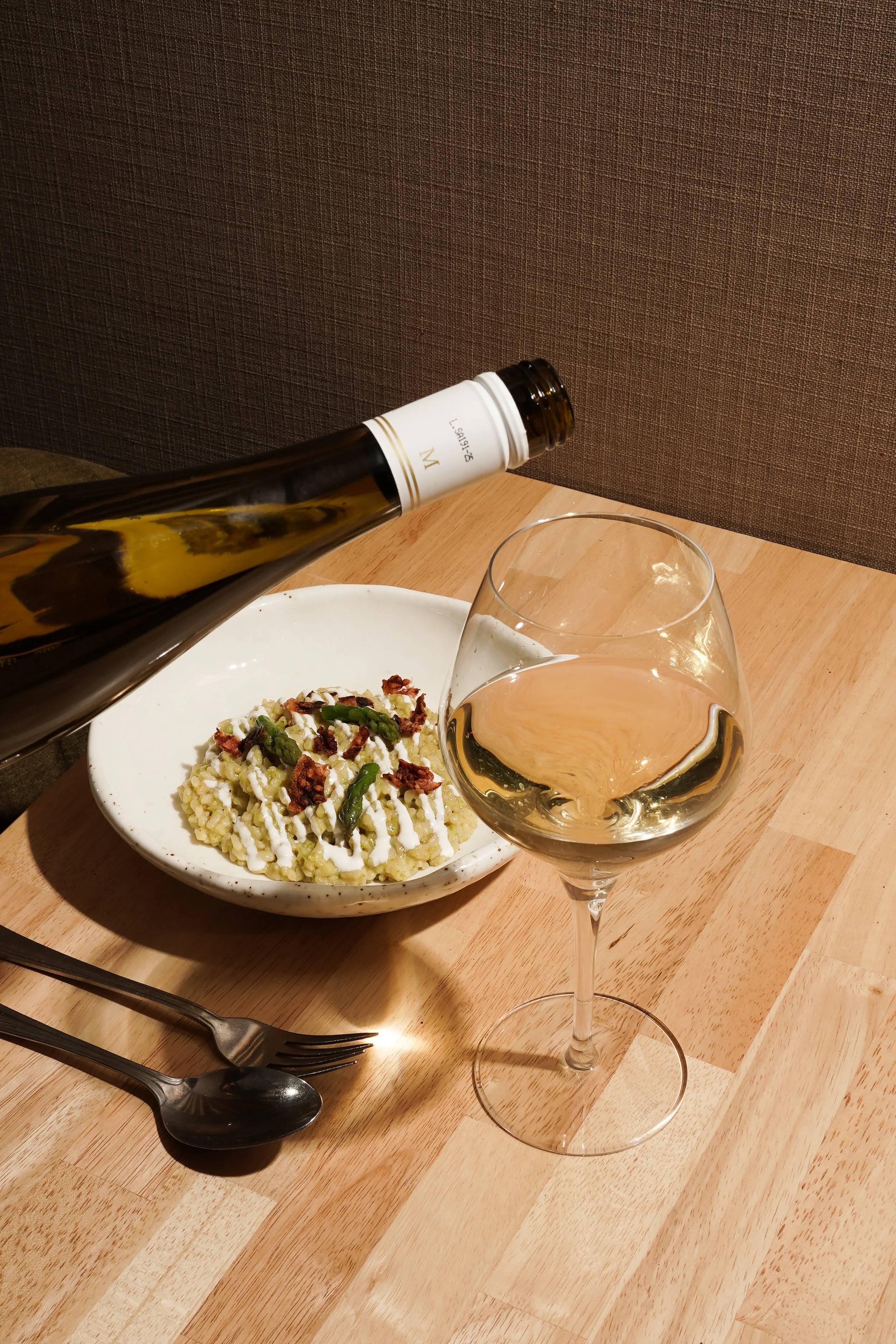 Verre de vin blanc, plat de risotto avec de la viande et des asperges, fourchette et cuillère sur une table en bois.