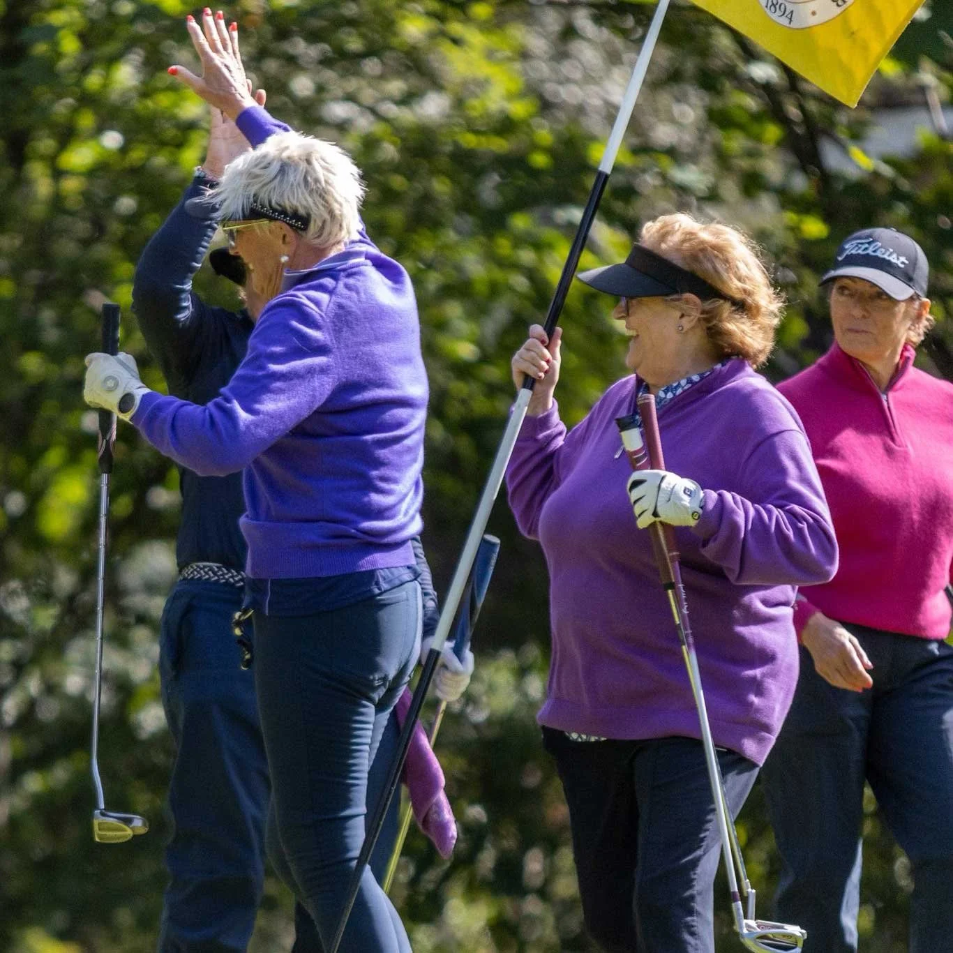 Three women with golf clubs in a park, dressed in colorful athletic clothing, participating in a golf activity.