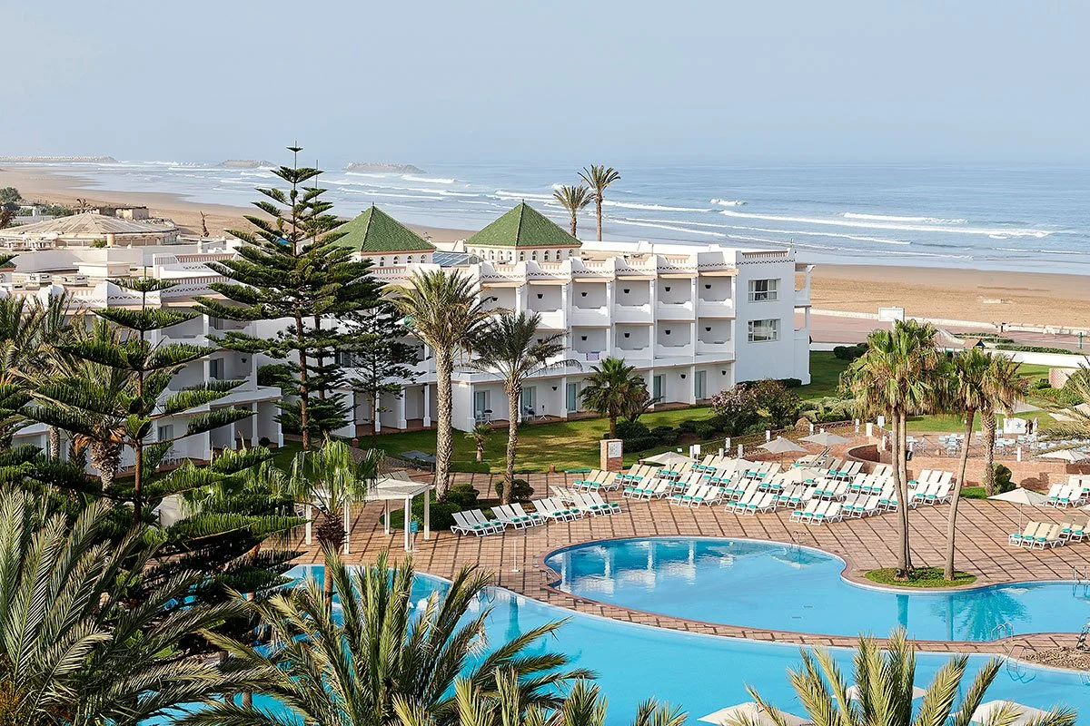 A beachfront resort with a large swimming pool, surrounded by lounge chairs and palm trees, overlooking the ocean.