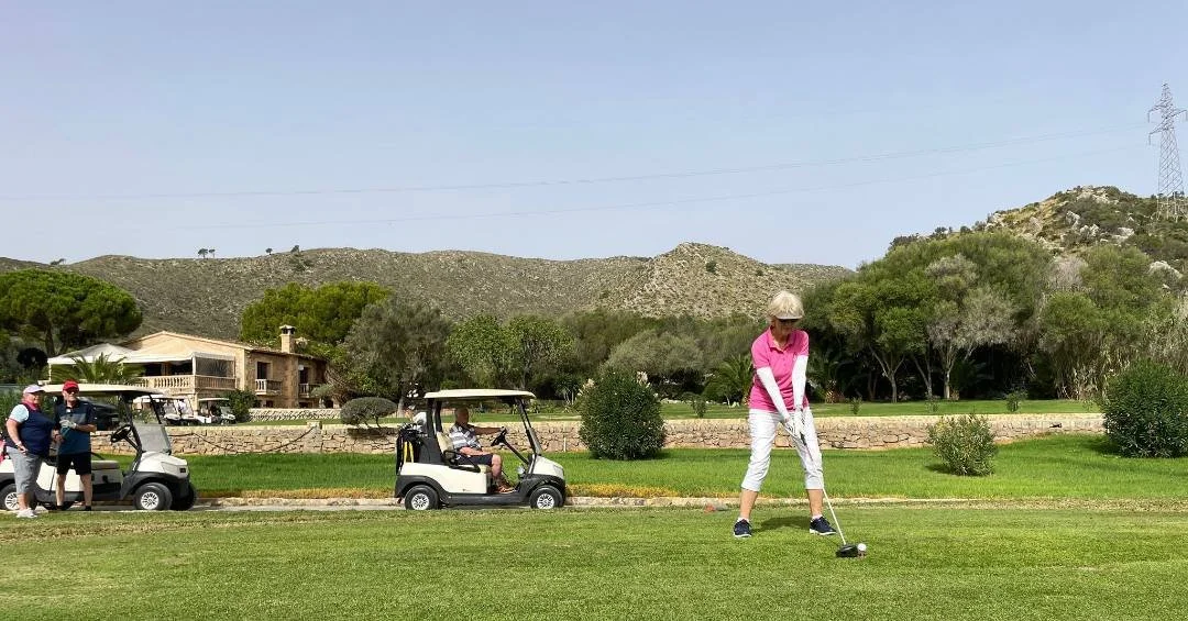 A woman in a pink shirt and white pants prepares to tee off on a golf course. In the background, two men stand near golf carts, and another person is sitting in a golf cart. The setting includes green grass, trees, a stone wall, and hills under a clear blue sky.