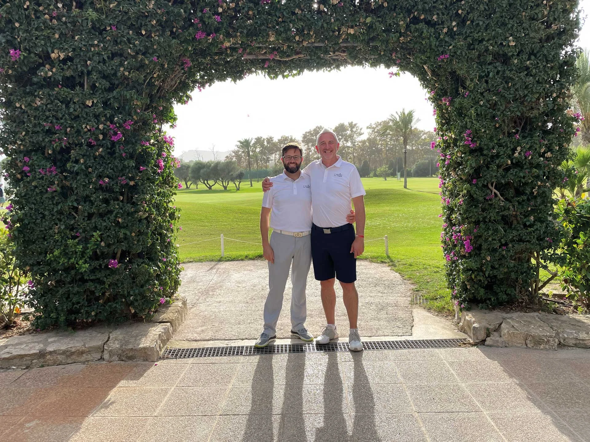 Two men standing under a floral arch on a golf course, smiling, with trees and palm trees in the background, during daytime.
