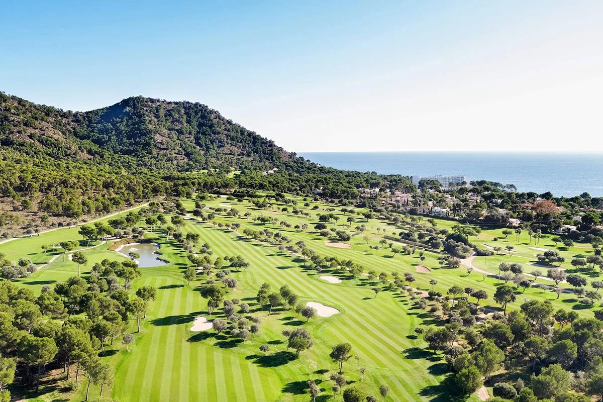 A scenic view of a golf course near the coast with lush green fairways, scattered trees, water hazards, and a mountain in the background