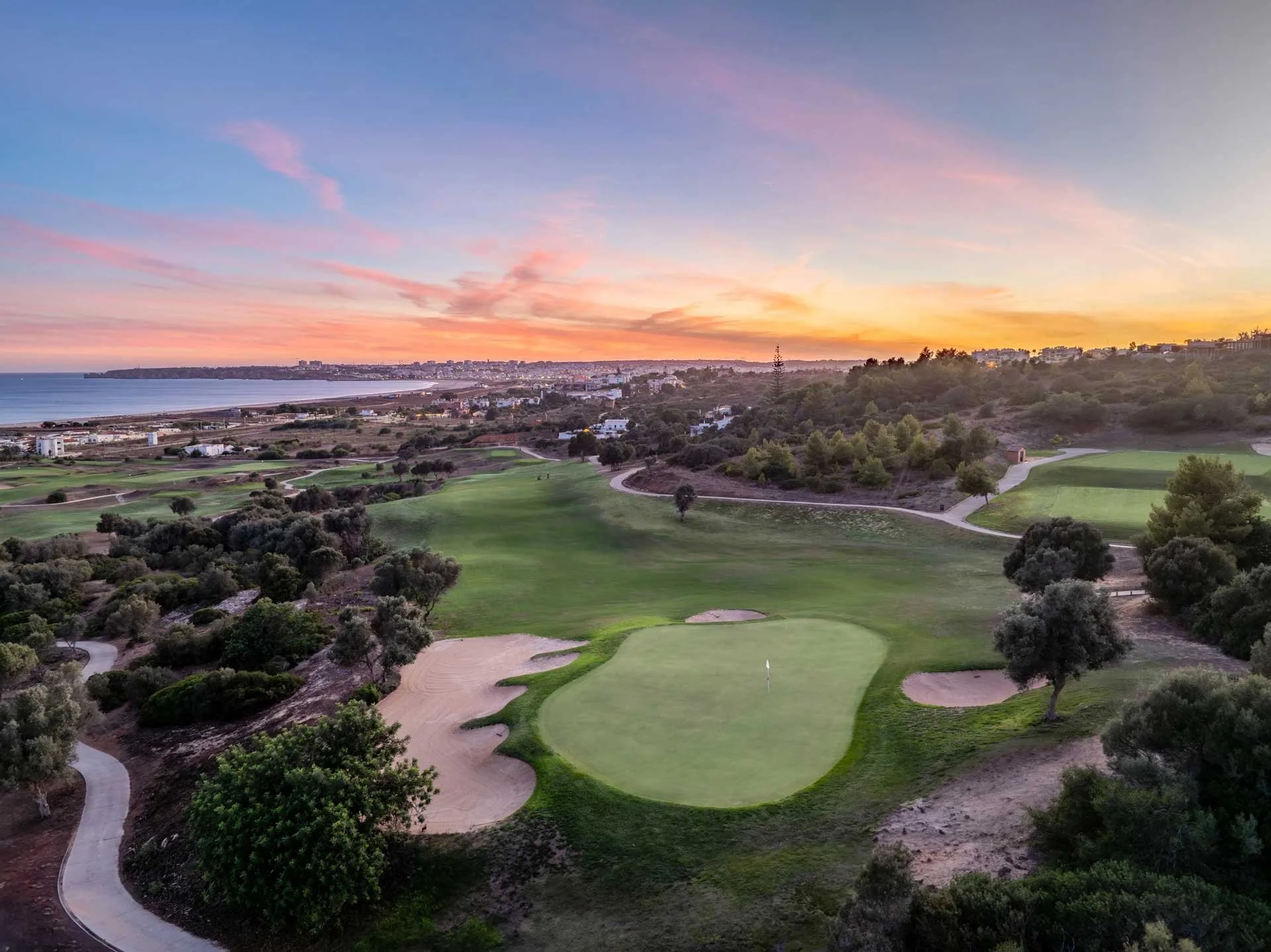 A scenic view of a golf course at sunset with a green field, sand traps, trees, and a distant cityscape along the coastline under a colorful sky with pink, orange, and purple clouds.