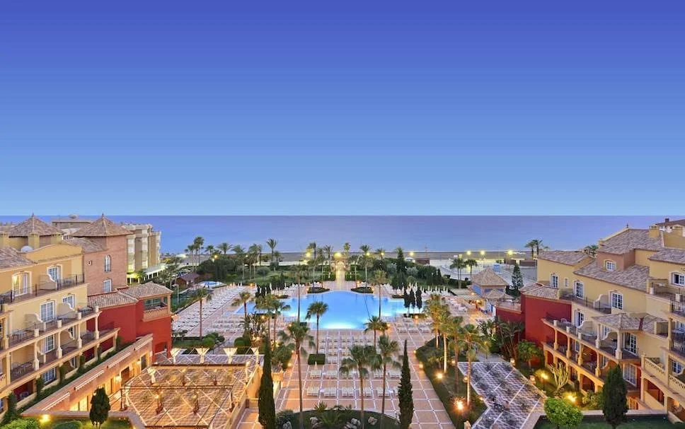 A view of a resort with a large swimming pool, surrounded by palm trees and lounge areas, with hotel buildings on either side, overlooking the ocean under a clear sky.