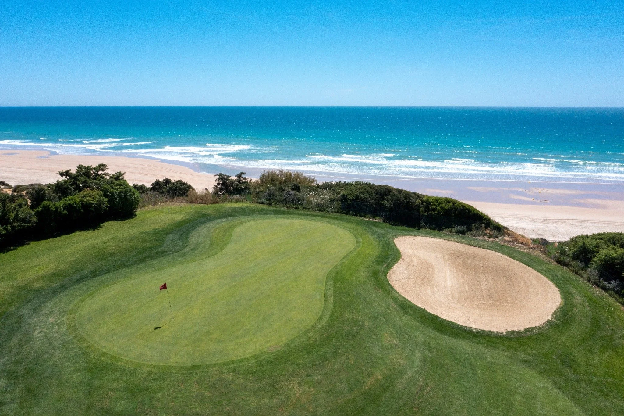 A scenic view of a golf course next to a beach with blue ocean water and clear sky.