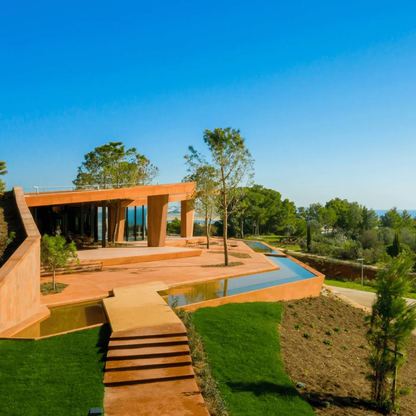 Modern house with glass walls, surrounded by trees, with a terraced landscape, water features, and a grassy lawn under a clear blue sky.